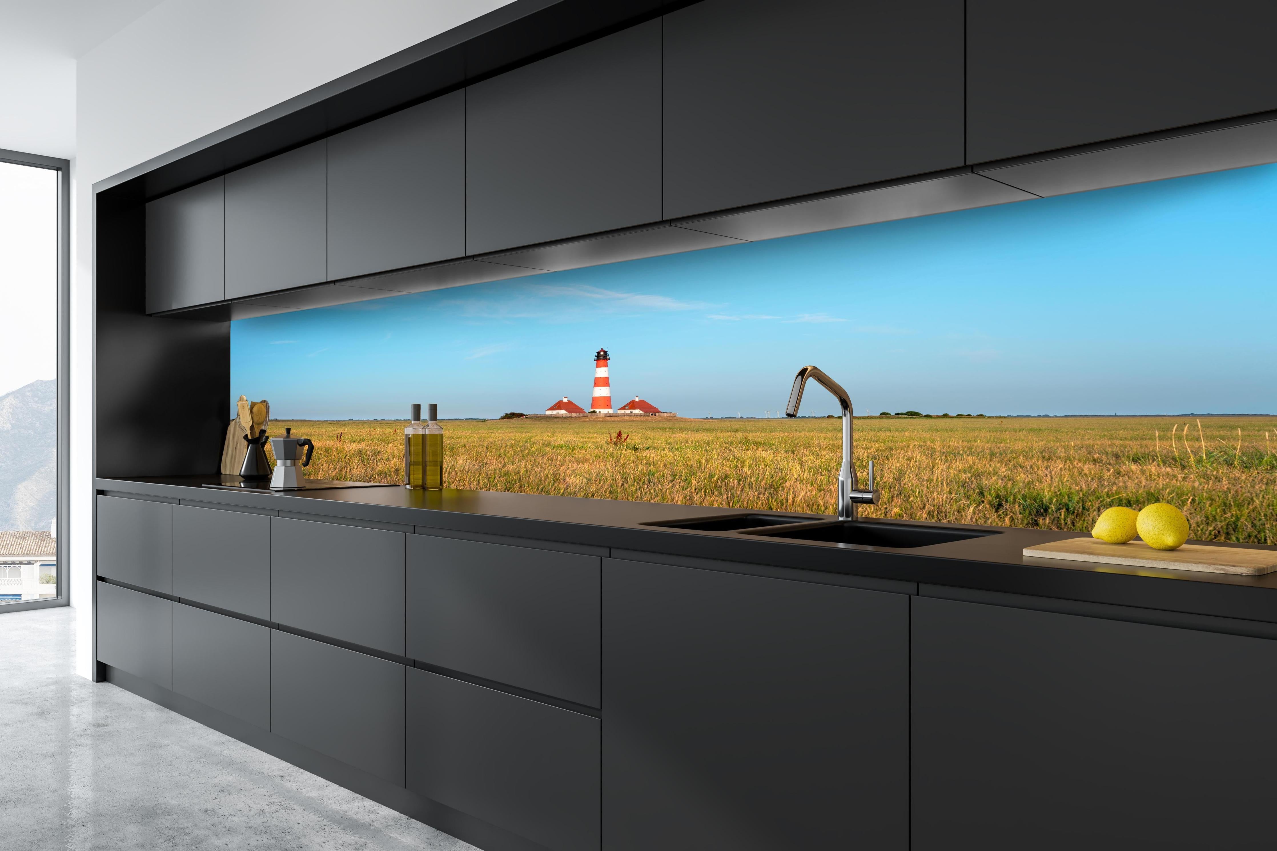 Küche - Ein beeindruckender Leuchtturm in der weitläufigen norddeutschen Landschaft unter blauem Himmel. hinter weißen Hochglanz-Küchenregalen und schwarzem Wasserhahn