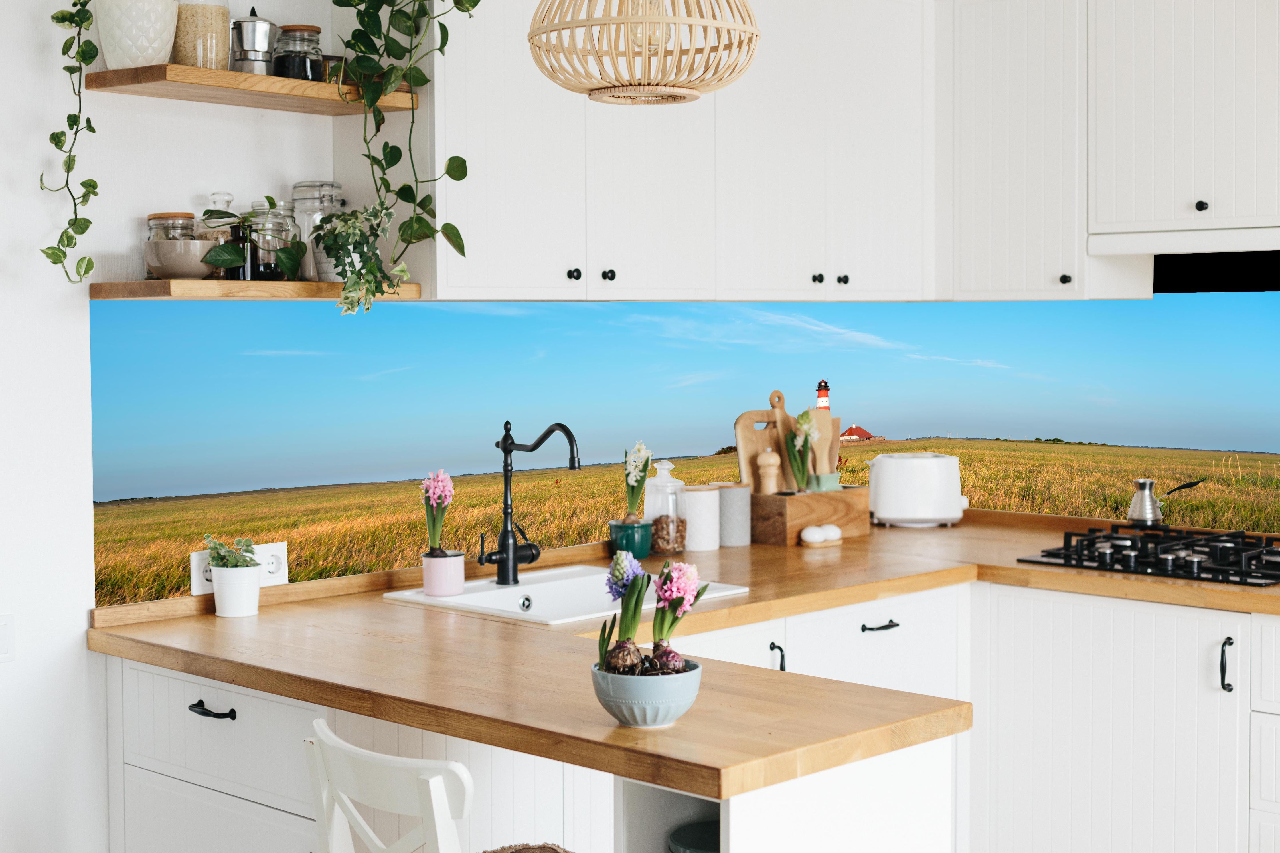 Küche - Ein beeindruckender Leuchtturm in der weitläufigen norddeutschen Landschaft unter blauem Himmel. hinter weißen Hochglanz-Küchenregalen und schwarzem Wasserhahn