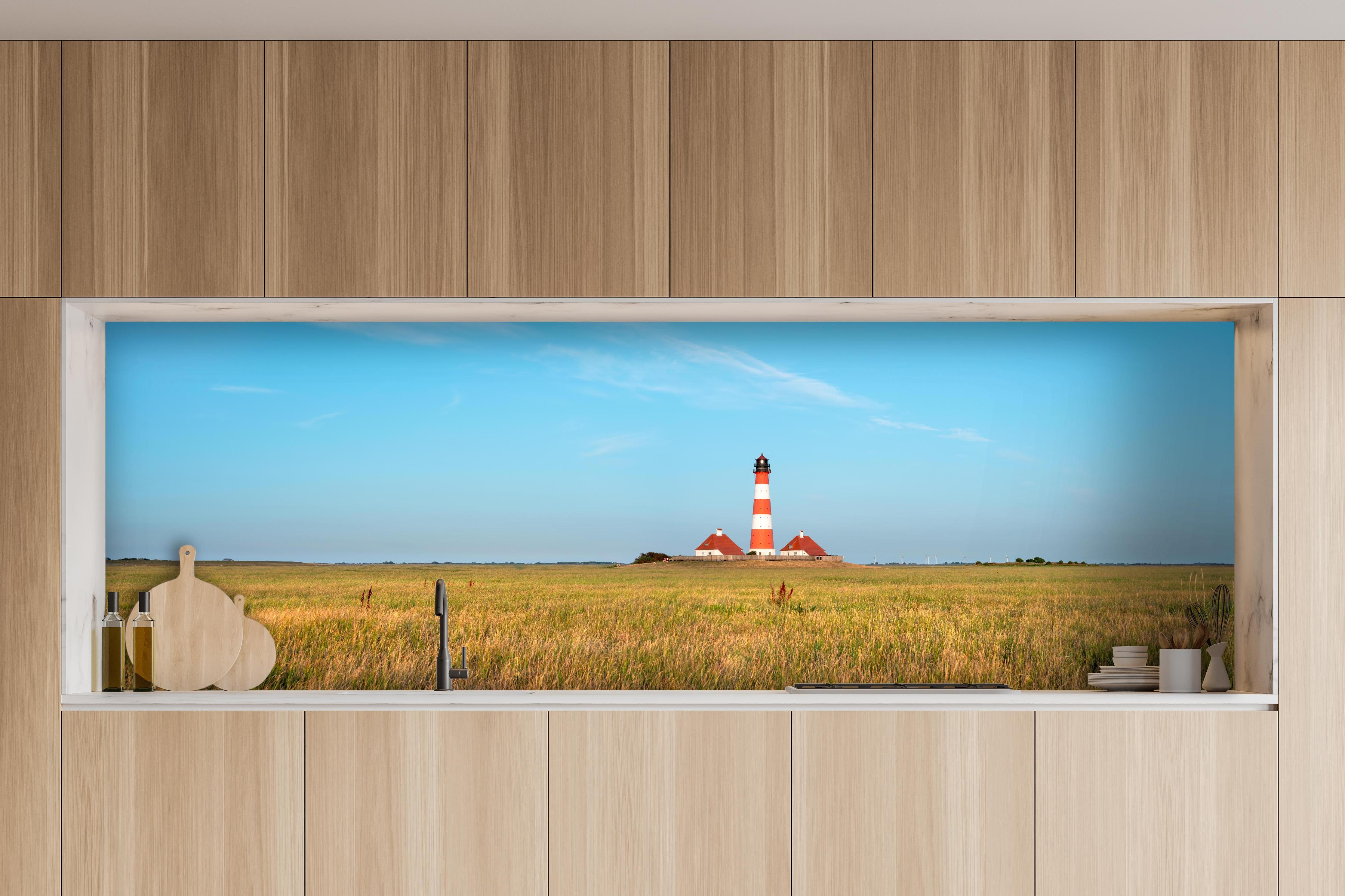 Küche - Ein beeindruckender Leuchtturm in der weitläufigen norddeutschen Landschaft unter blauem Himmel. hinter weißen Hochglanz-Küchenregalen und schwarzem Wasserhahn