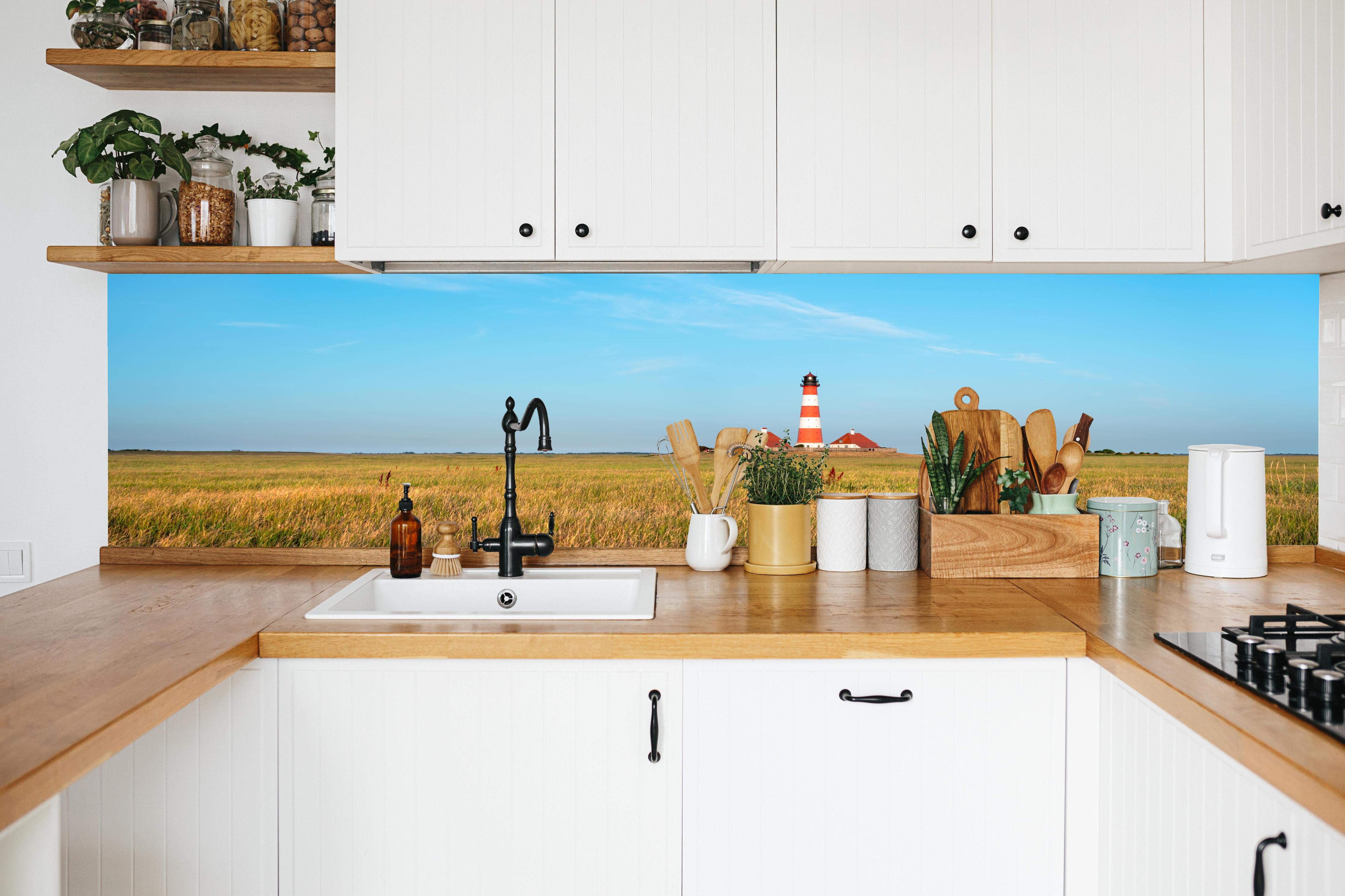 Küche - Ein beeindruckender Leuchtturm in der weitläufigen norddeutschen Landschaft unter blauem Himmel. hinter weißen Hochglanz-Küchenregalen und schwarzem Wasserhahn