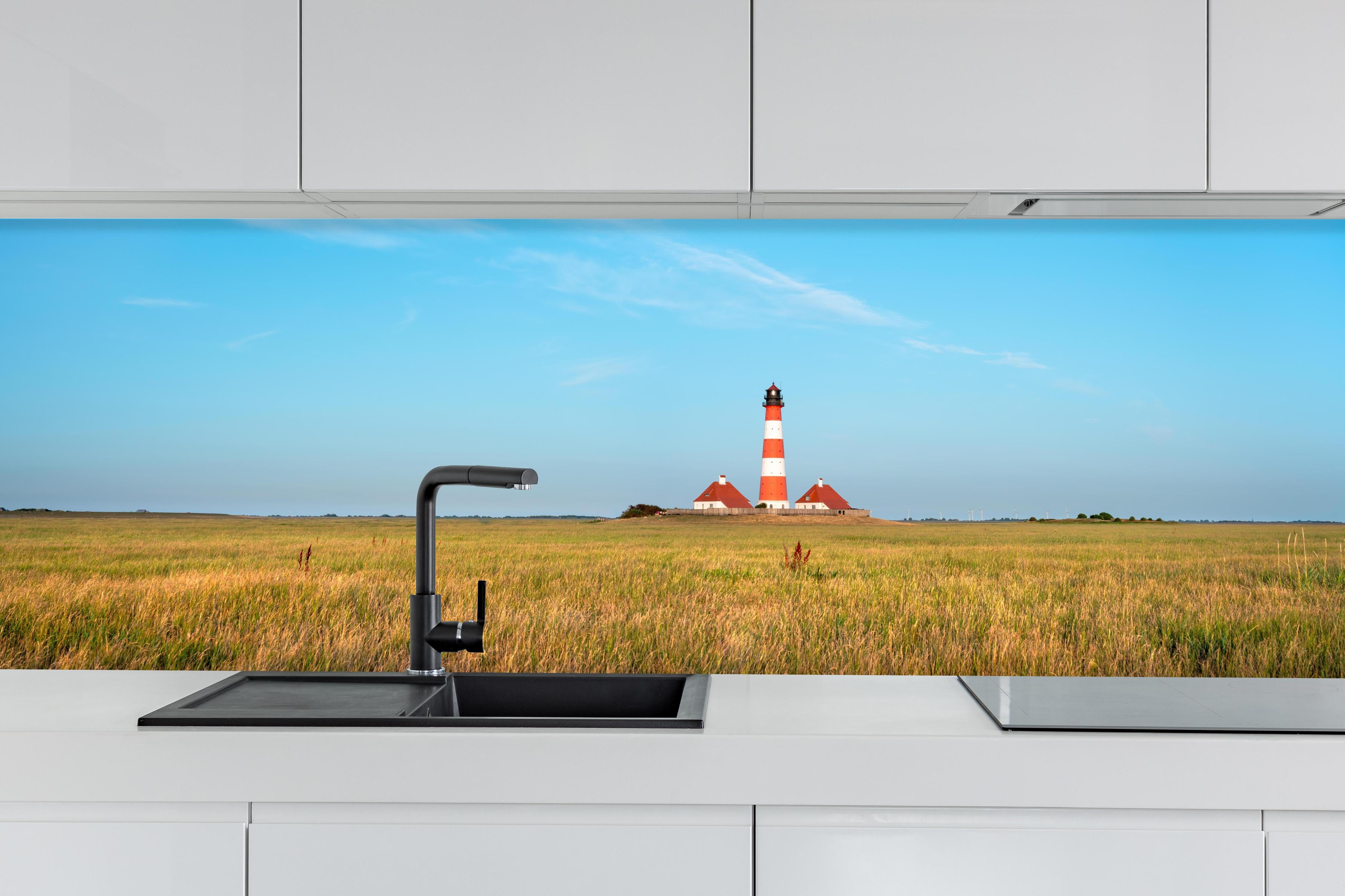 Küche - Ein beeindruckender Leuchtturm in der weitläufigen norddeutschen Landschaft unter blauem Himmel. hinter weißen Hochglanz-Küchenregalen und schwarzem Wasserhahn