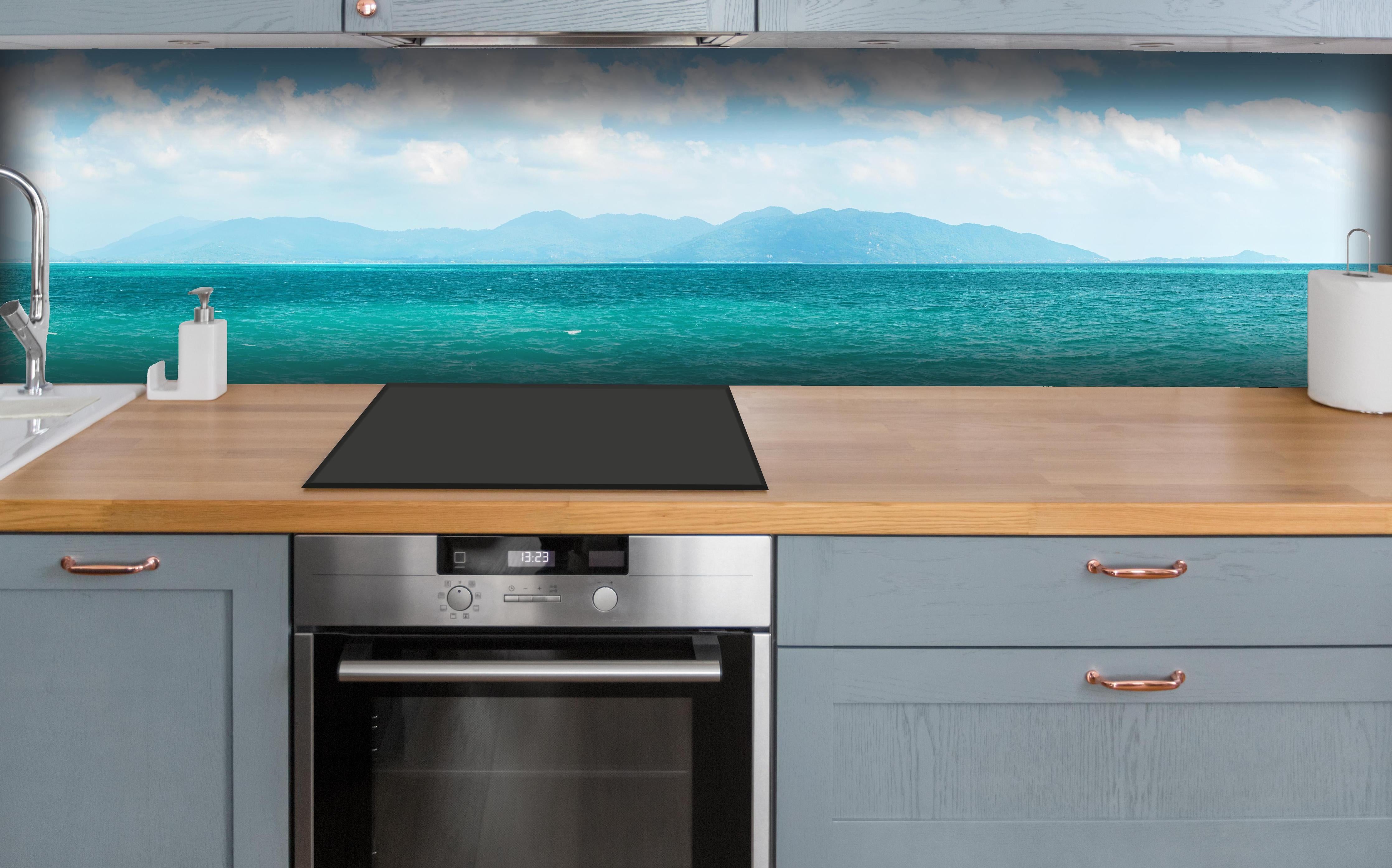 Küche - Ein beeindruckendes Panorama des Ozeans unter einem klaren blauen Himmel mit sanften Wellen und malerischen Bergen im Hintergrund. hinter weißen Hochglanz-Küchenregalen und schwarzem Wasserhahn