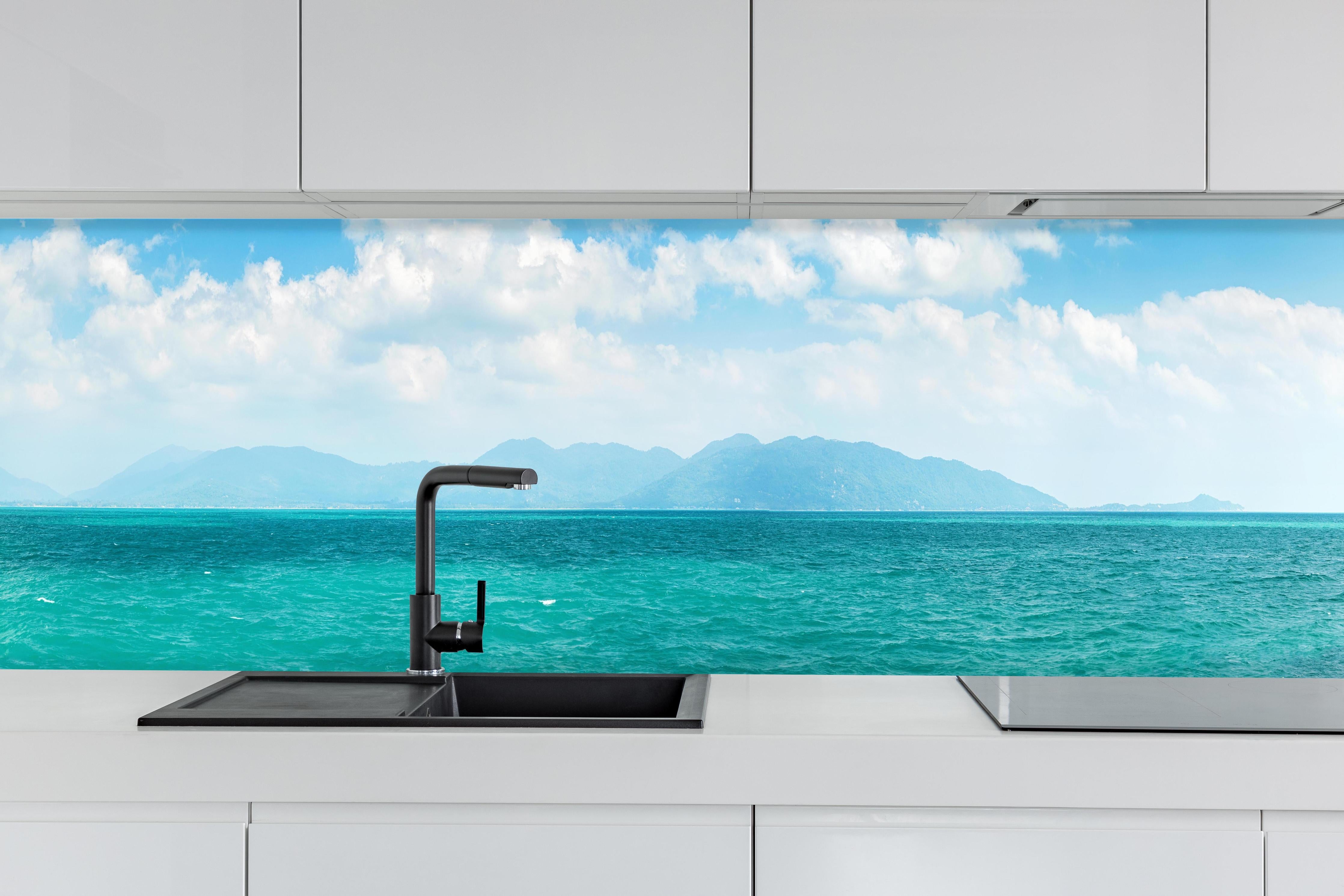Küche - Ein beeindruckendes Panorama des Ozeans unter einem klaren blauen Himmel mit sanften Wellen und malerischen Bergen im Hintergrund. hinter weißen Hochglanz-Küchenregalen und schwarzem Wasserhahn