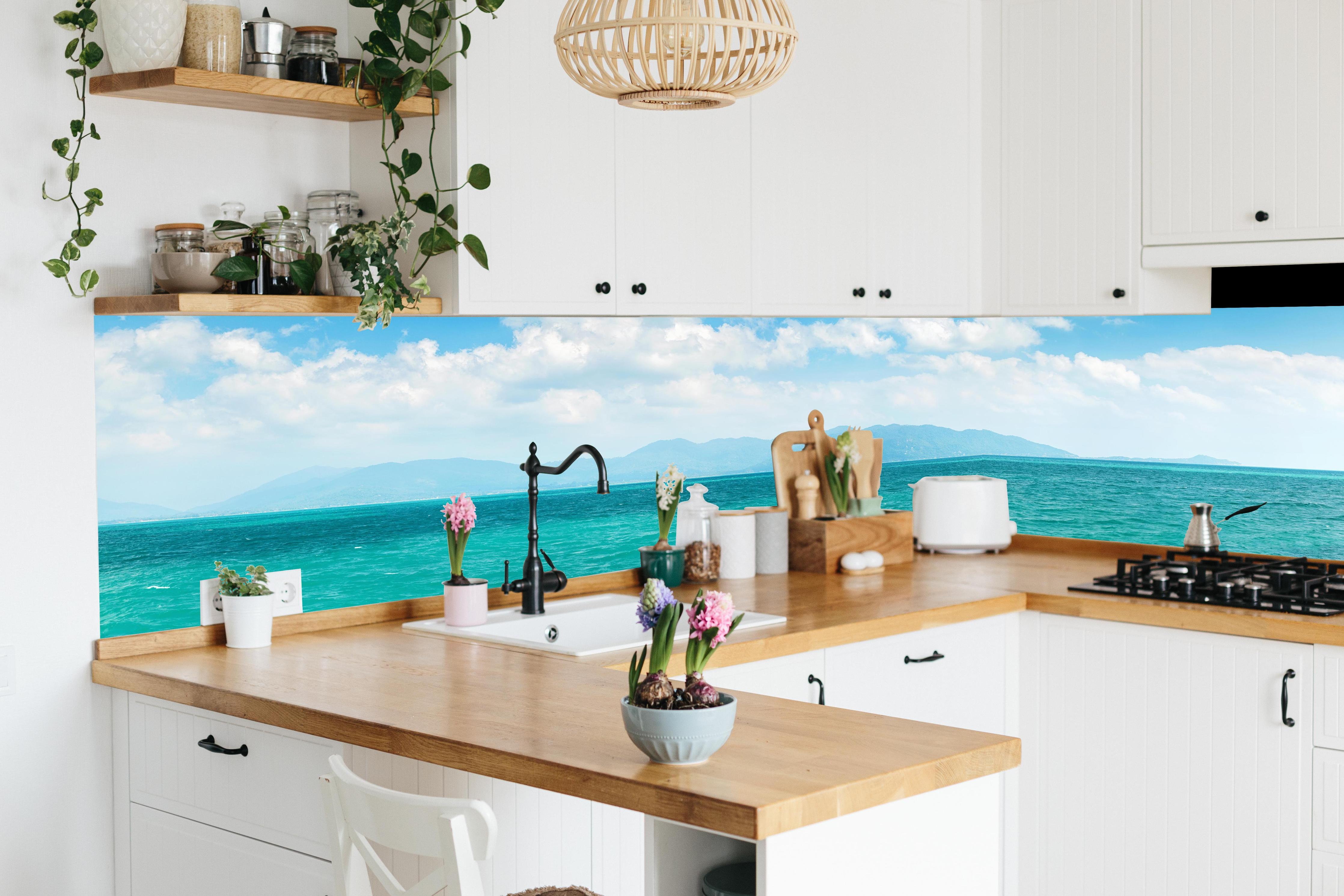 Küche - Ein beeindruckendes Panorama des Ozeans unter einem klaren blauen Himmel mit sanften Wellen und malerischen Bergen im Hintergrund. hinter weißen Hochglanz-Küchenregalen und schwarzem Wasserhahn