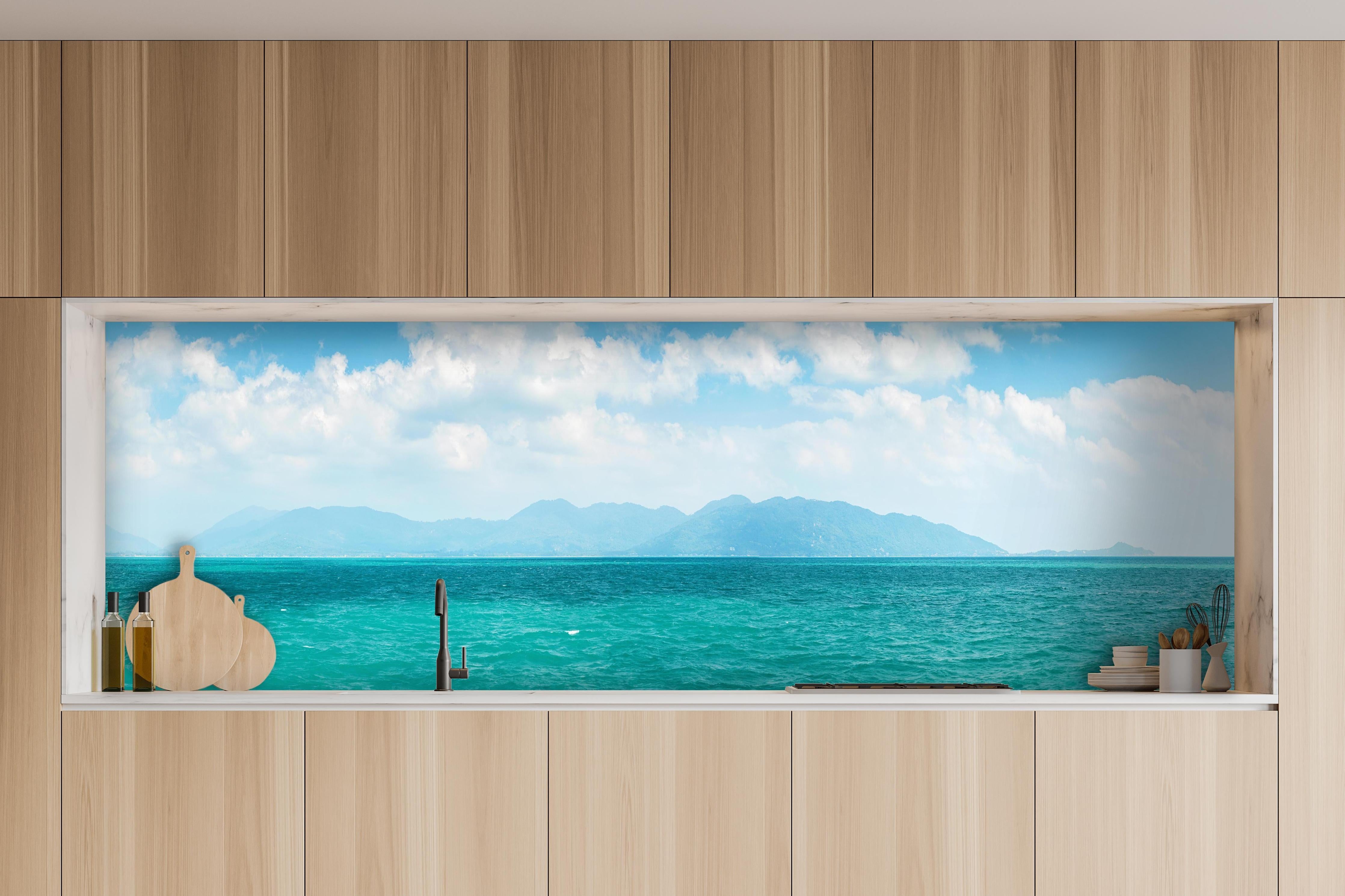 Küche - Ein beeindruckendes Panorama des Ozeans unter einem klaren blauen Himmel mit sanften Wellen und malerischen Bergen im Hintergrund. hinter weißen Hochglanz-Küchenregalen und schwarzem Wasserhahn