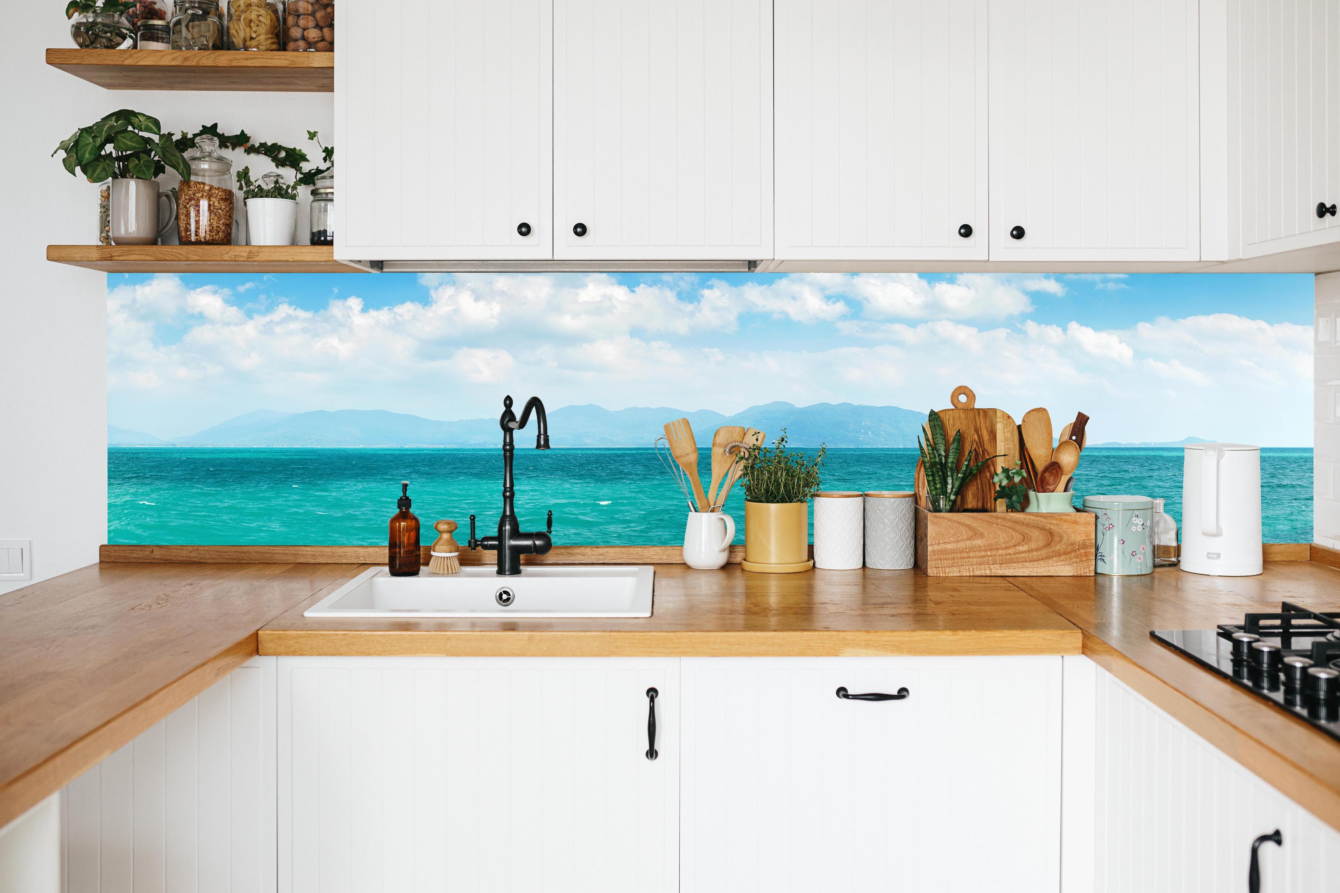 Küche - Ein beeindruckendes Panorama des Ozeans unter einem klaren blauen Himmel mit sanften Wellen und malerischen Bergen im Hintergrund. hinter weißen Hochglanz-Küchenregalen und schwarzem Wasserhahn