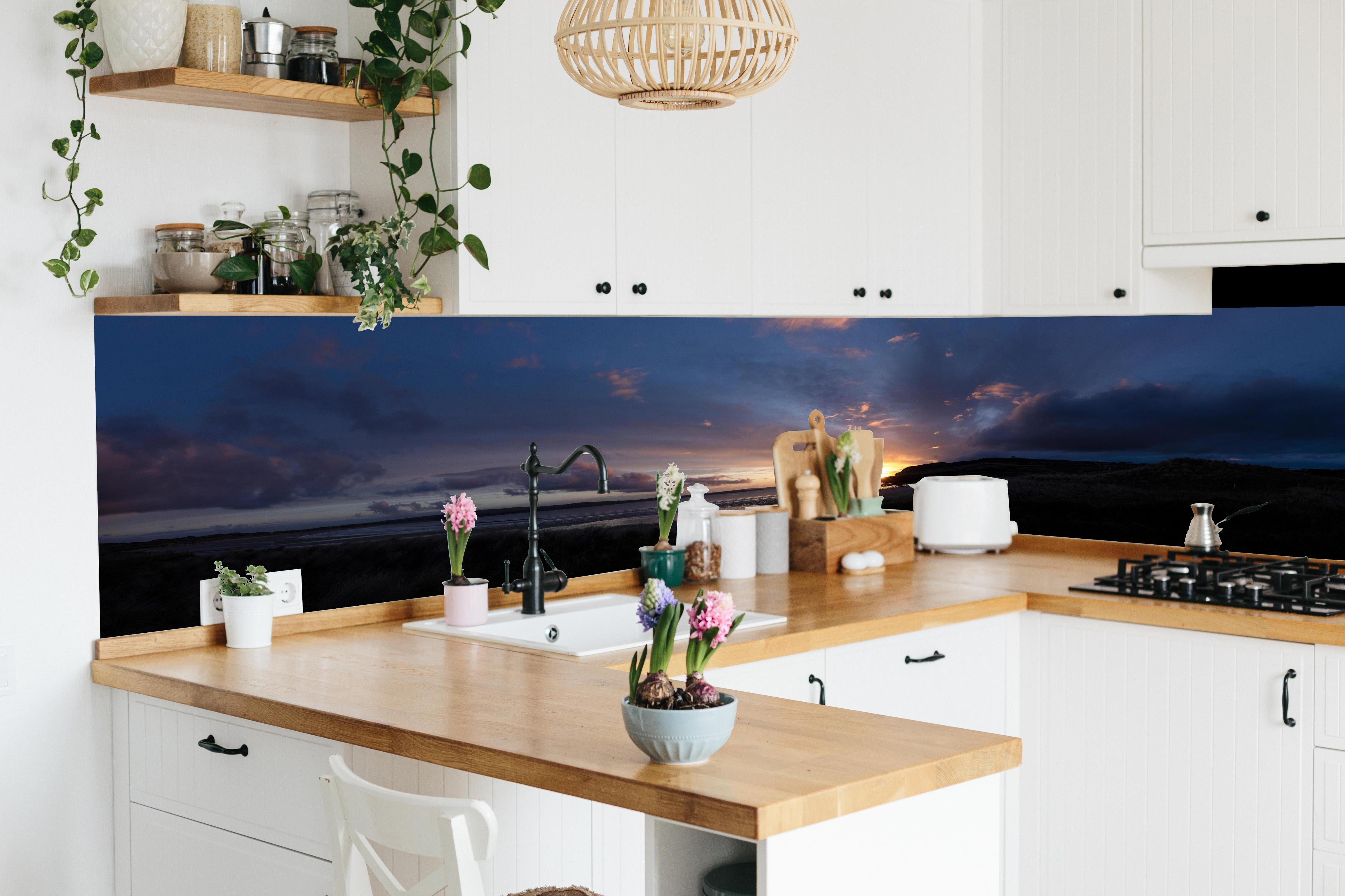 Küche - Ein malerischer Sonnenuntergang über einem ruhigen See, die Wolken leuchten in verschiedenen Blau- und Orangetönen hinter weißen Hochglanz-Küchenregalen und schwarzem Wasserhahn