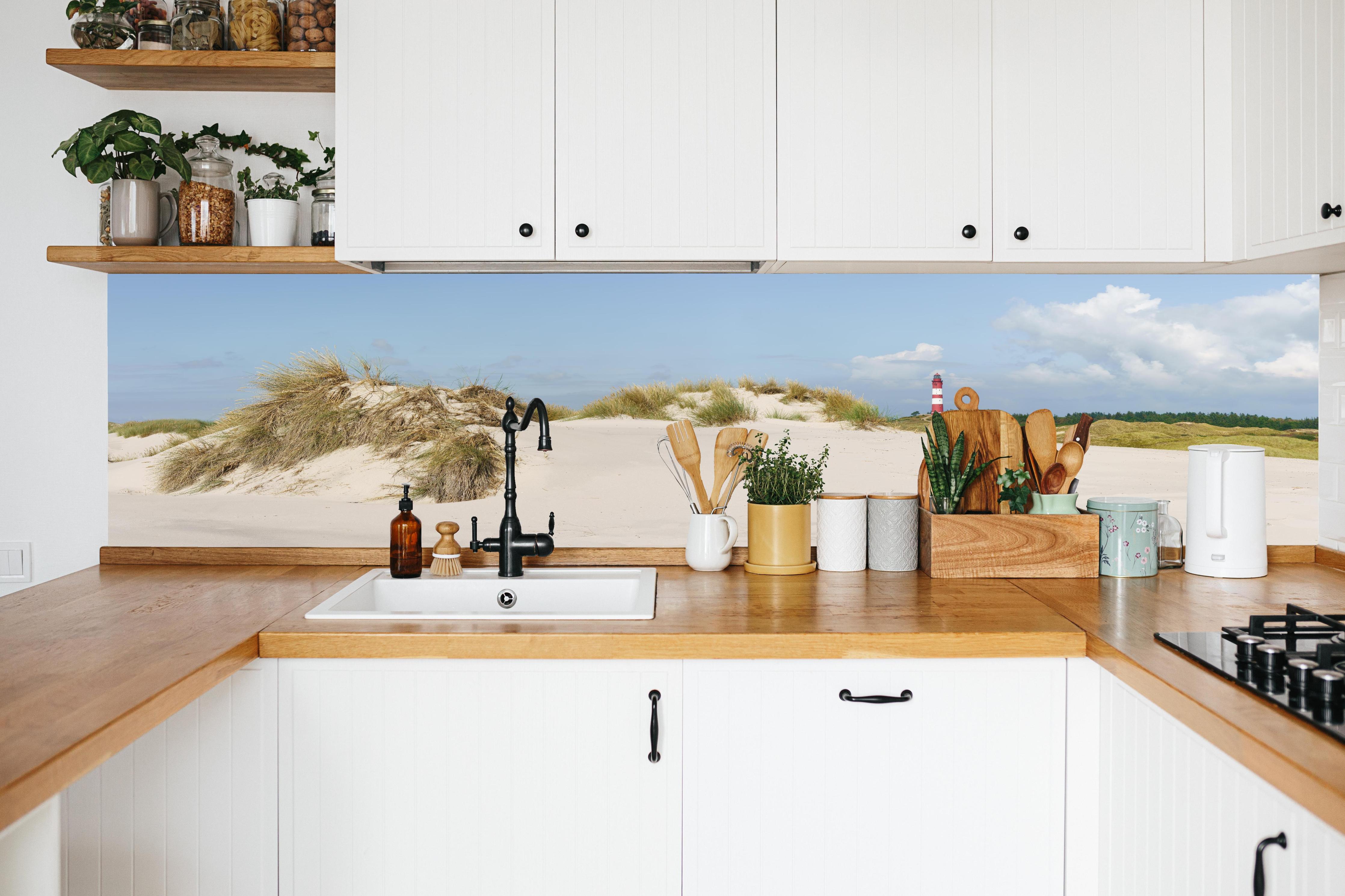 Küche - Ein traumhafter Blick auf die weiße Sanddüne und den roten Leuchtturm am Meer hinter weißen Hochglanz-Küchenregalen und schwarzem Wasserhahn