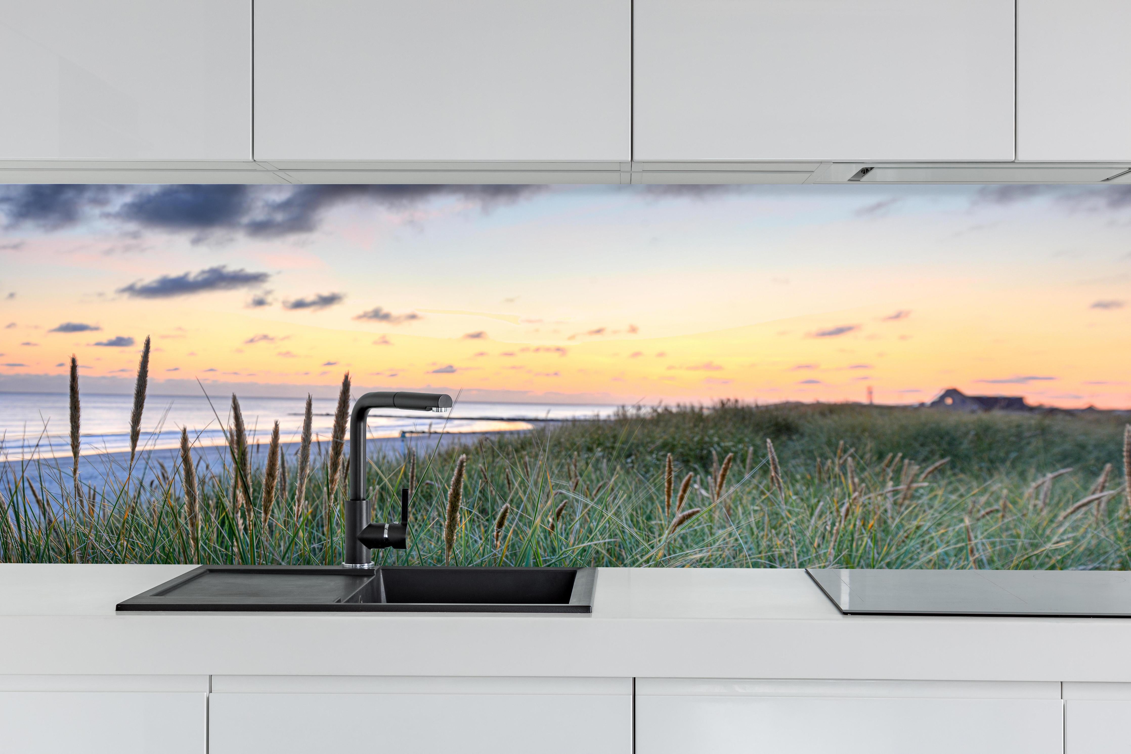 Küche - Friedliche Strandlandschaft mit grünem Gras und sanften Wellen bei Sonnenuntergang hinter weißen Hochglanz-Küchenregalen und schwarzem Wasserhahn