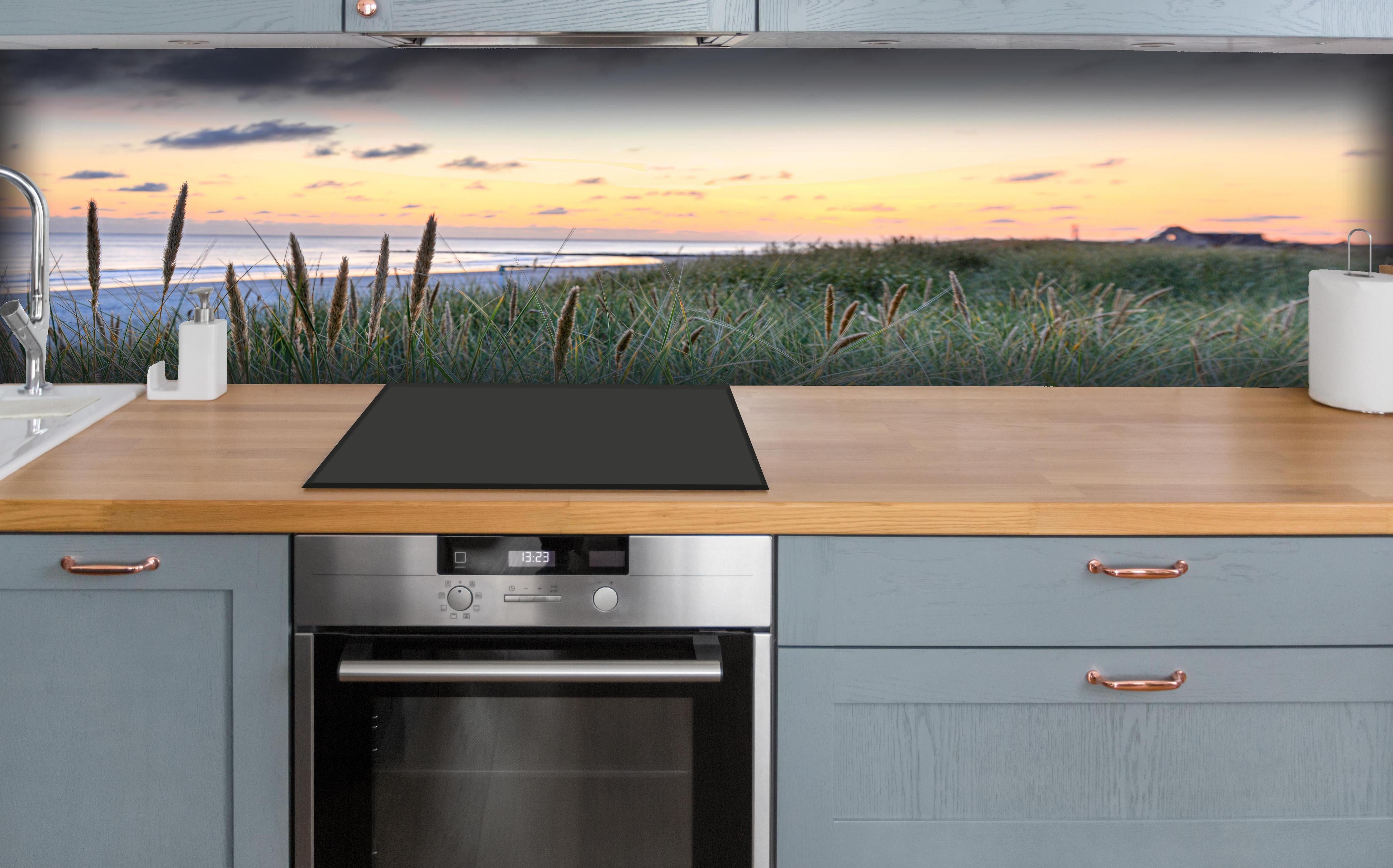 Küche - Friedliche Strandlandschaft mit grünem Gras und sanften Wellen bei Sonnenuntergang hinter weißen Hochglanz-Küchenregalen und schwarzem Wasserhahn