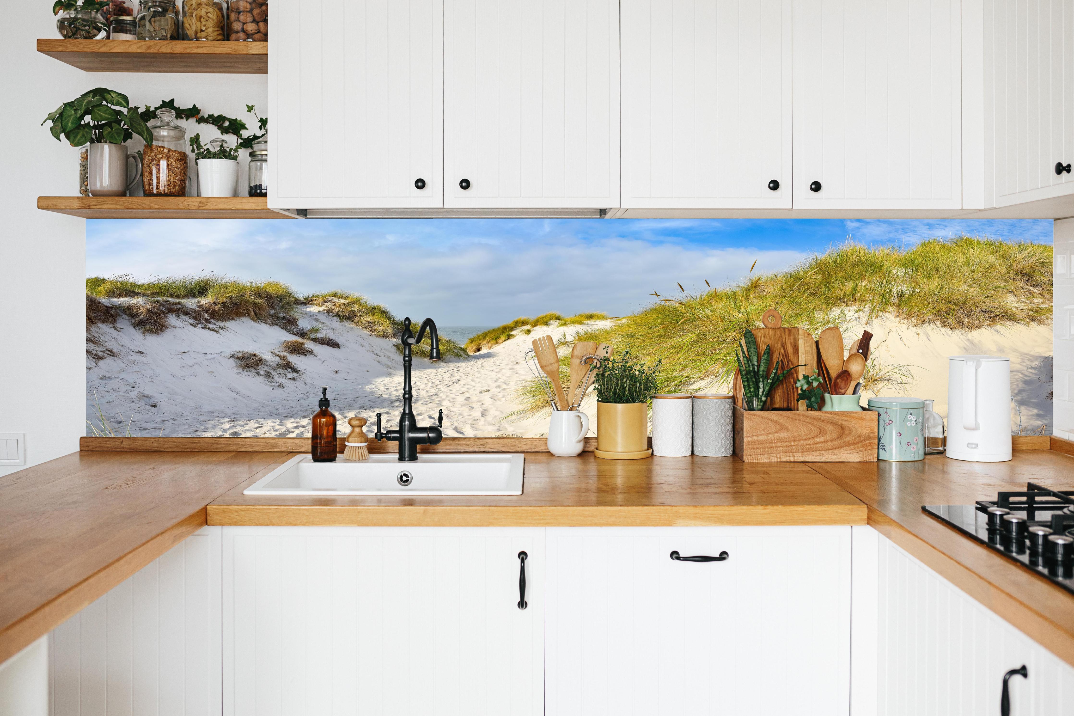 Küche - Idyllische Dünenlandschaft mit feinem Sand und grünem Gras hinter weißen Hochglanz-Küchenregalen und schwarzem Wasserhahn