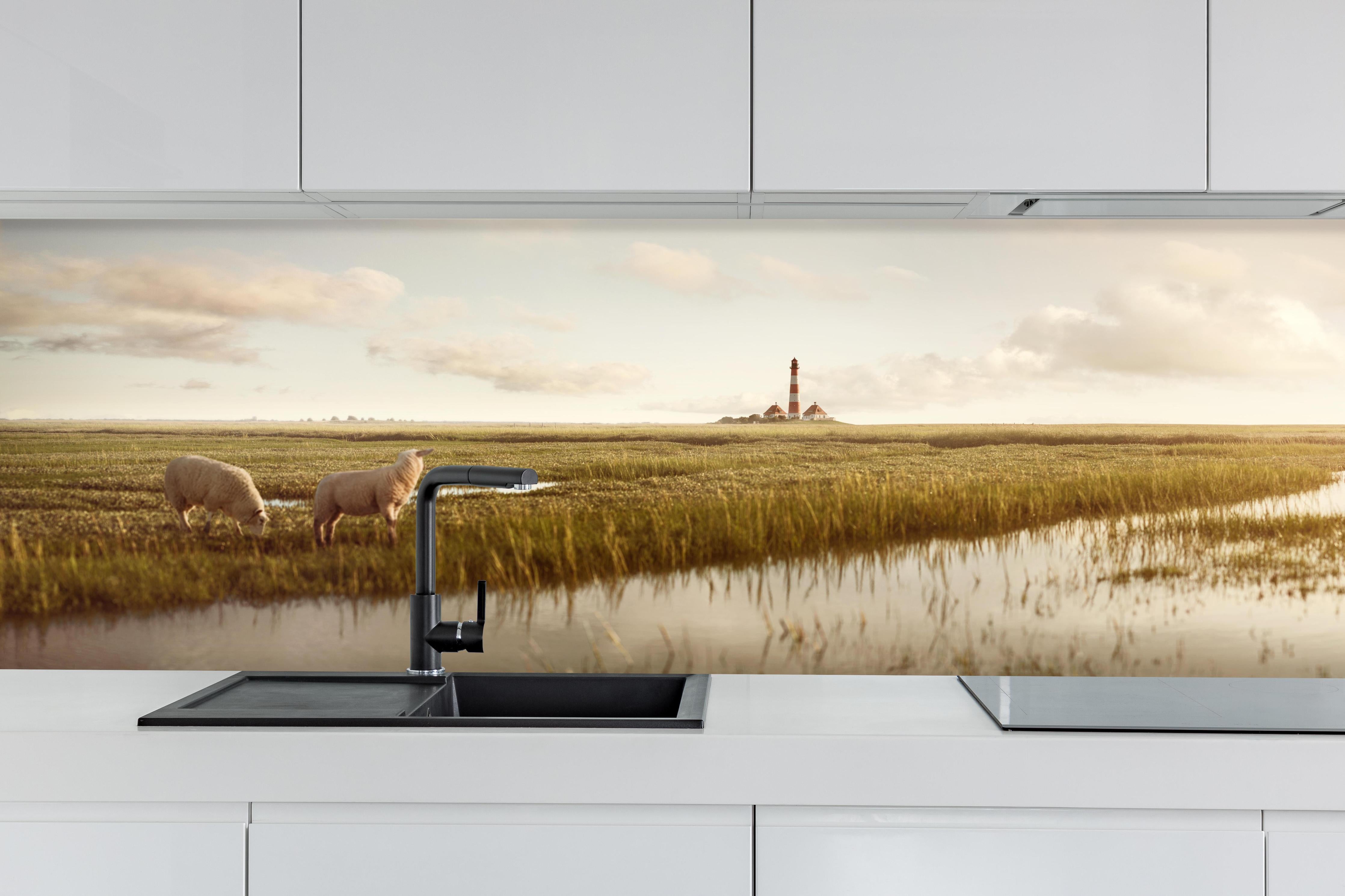 Küche - Idyllische Kulisse mit Schafen und einem Leuchtturm in ruhiger Küstenlandschaft hinter weißen Hochglanz-Küchenregalen und schwarzem Wasserhahn