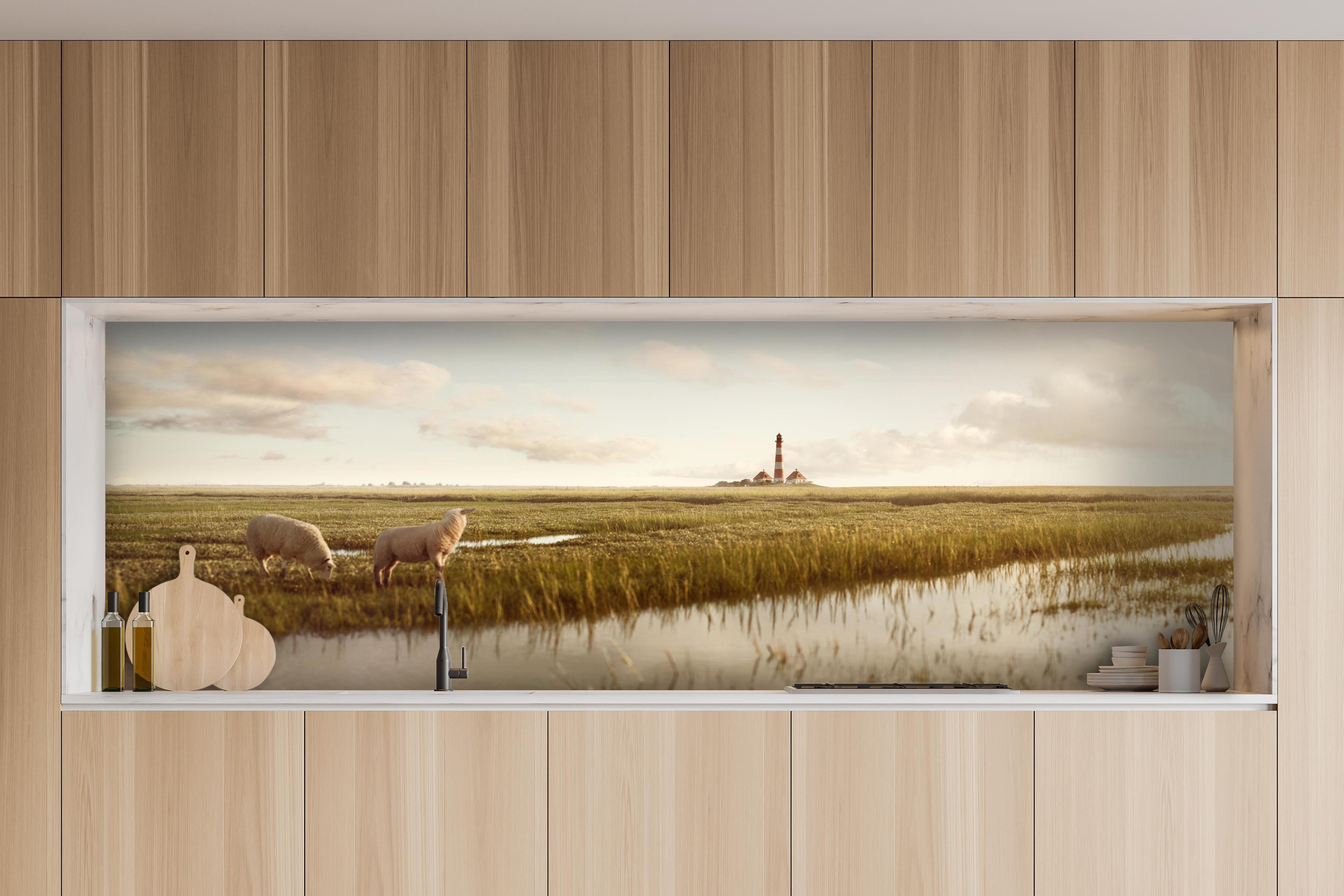 Küche - Idyllische Kulisse mit Schafen und einem Leuchtturm in ruhiger Küstenlandschaft hinter weißen Hochglanz-Küchenregalen und schwarzem Wasserhahn
