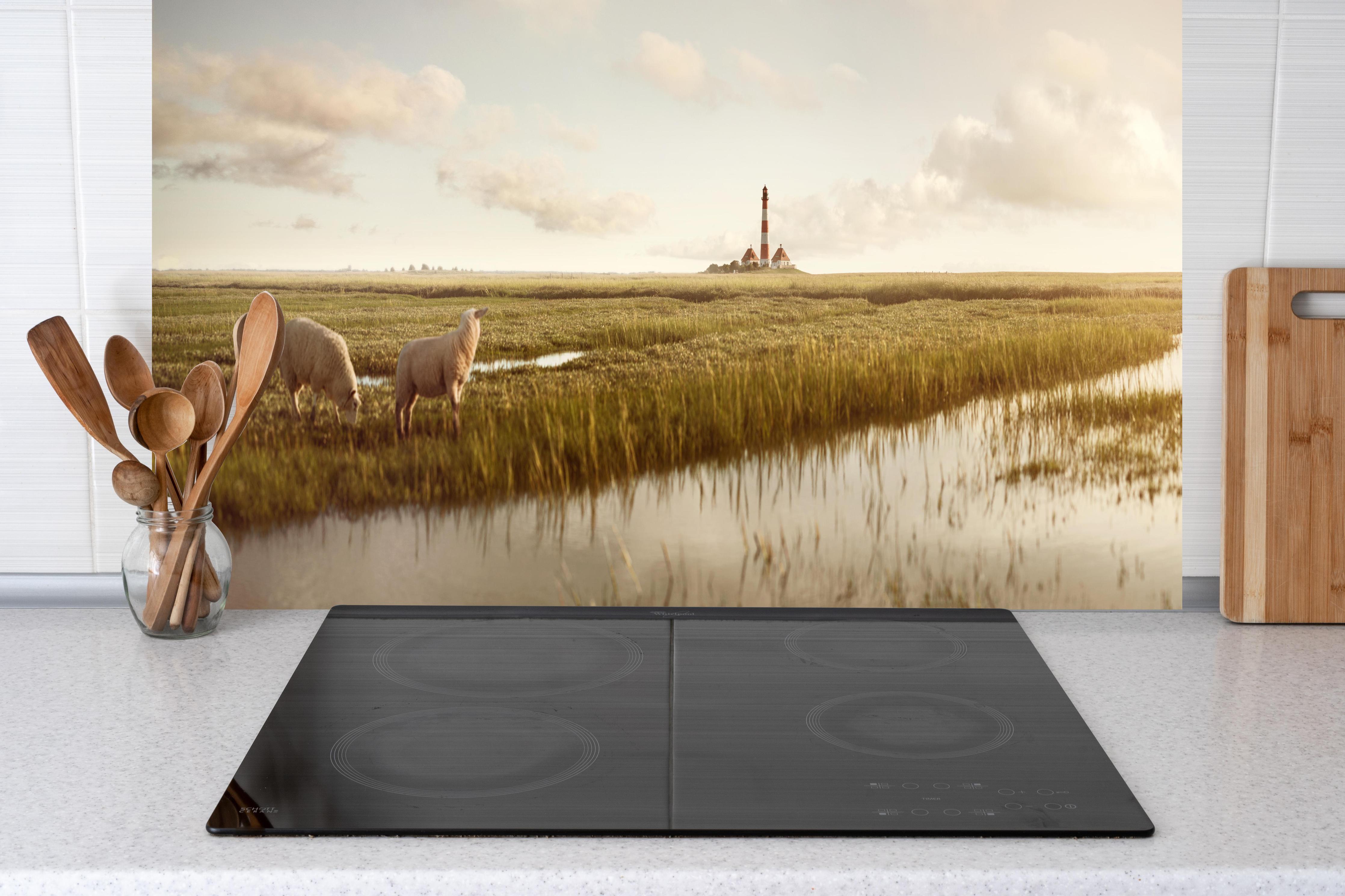 Küche - Idyllische Kulisse mit Schafen und einem Leuchtturm in ruhiger Küstenlandschaft hinter weißen Hochglanz-Küchenregalen und schwarzem Wasserhahn