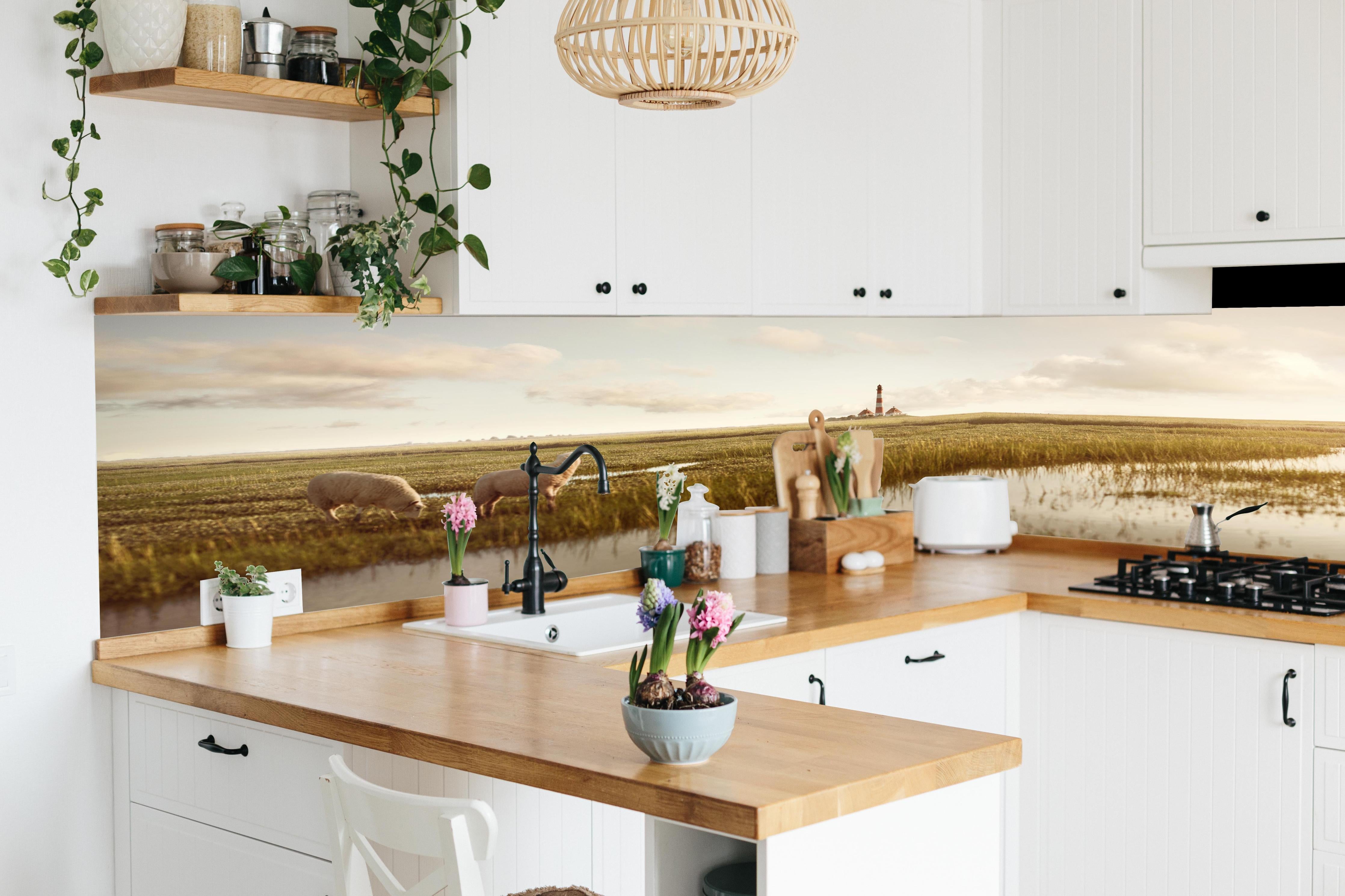 Küche - Idyllische Kulisse mit Schafen und einem Leuchtturm in ruhiger Küstenlandschaft hinter weißen Hochglanz-Küchenregalen und schwarzem Wasserhahn