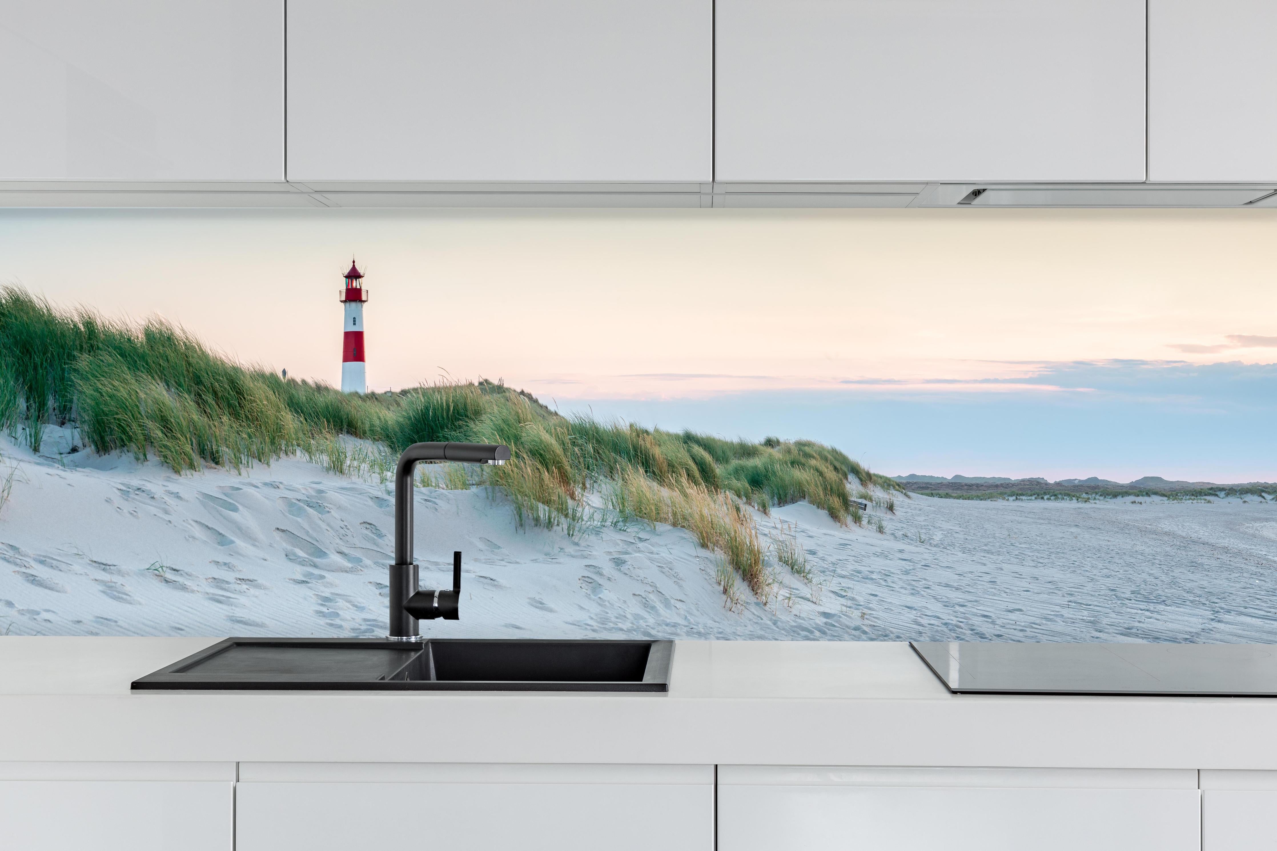Küche - Idyllischer Leuchtturm zwischen Sanddünen und sanften Wellen hinter weißen Hochglanz-Küchenregalen und schwarzem Wasserhahn