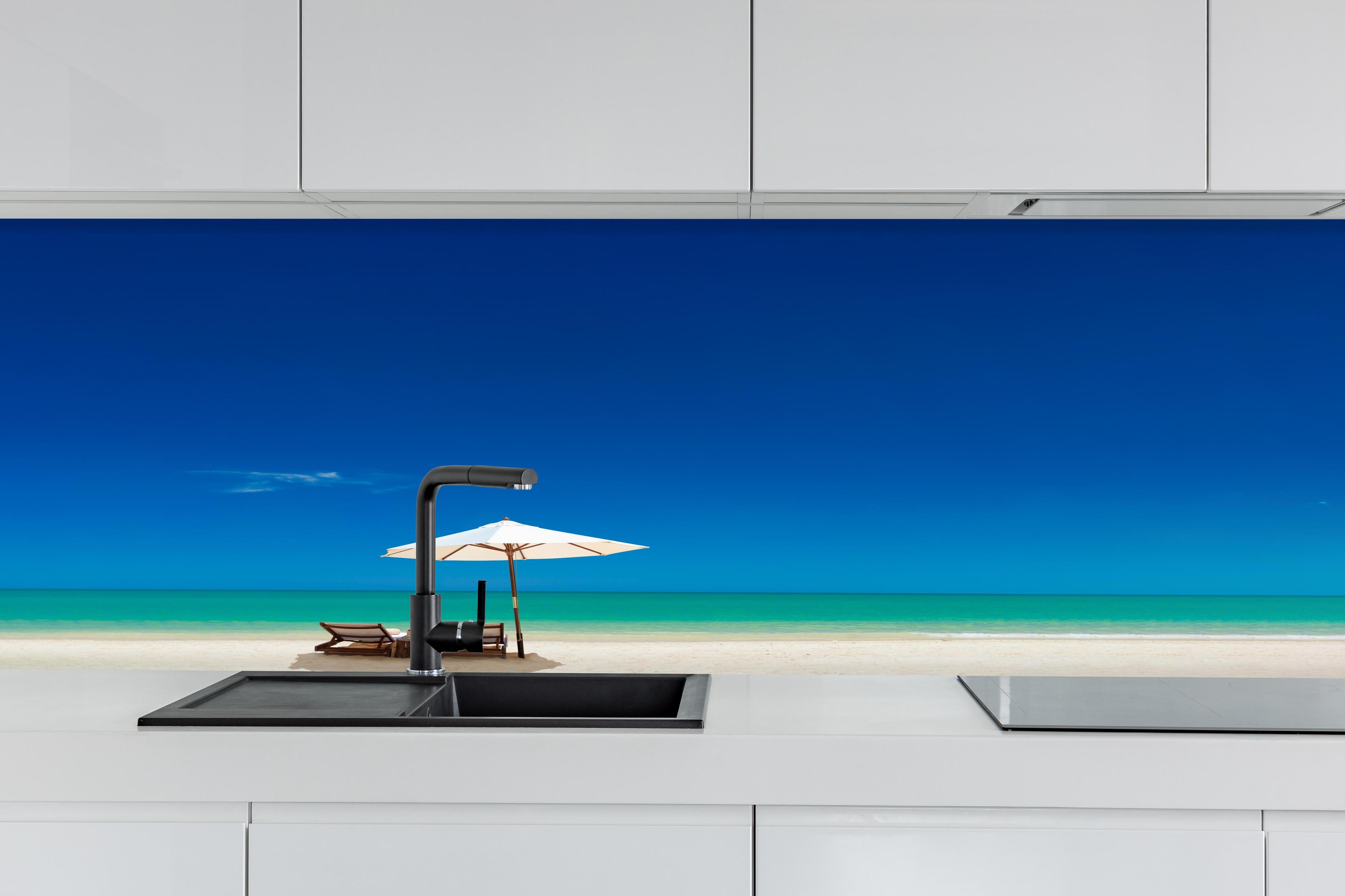 Küche - Idyllischer Strand mit Liegen und Sonnenschirm unter blauem Himmel hinter weißen Hochglanz-Küchenregalen und schwarzem Wasserhahn