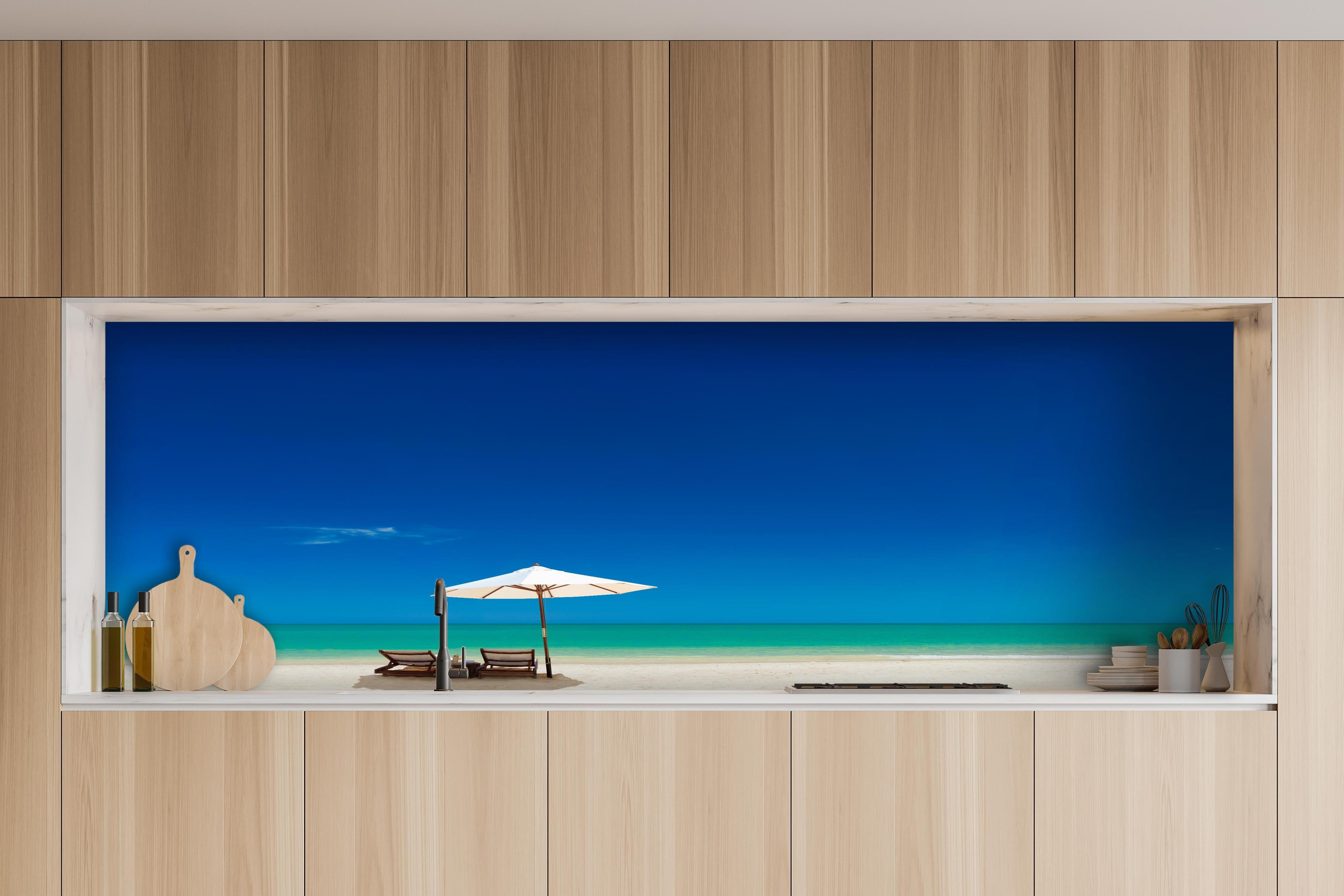 Küche - Idyllischer Strand mit Liegen und Sonnenschirm unter blauem Himmel hinter weißen Hochglanz-Küchenregalen und schwarzem Wasserhahn