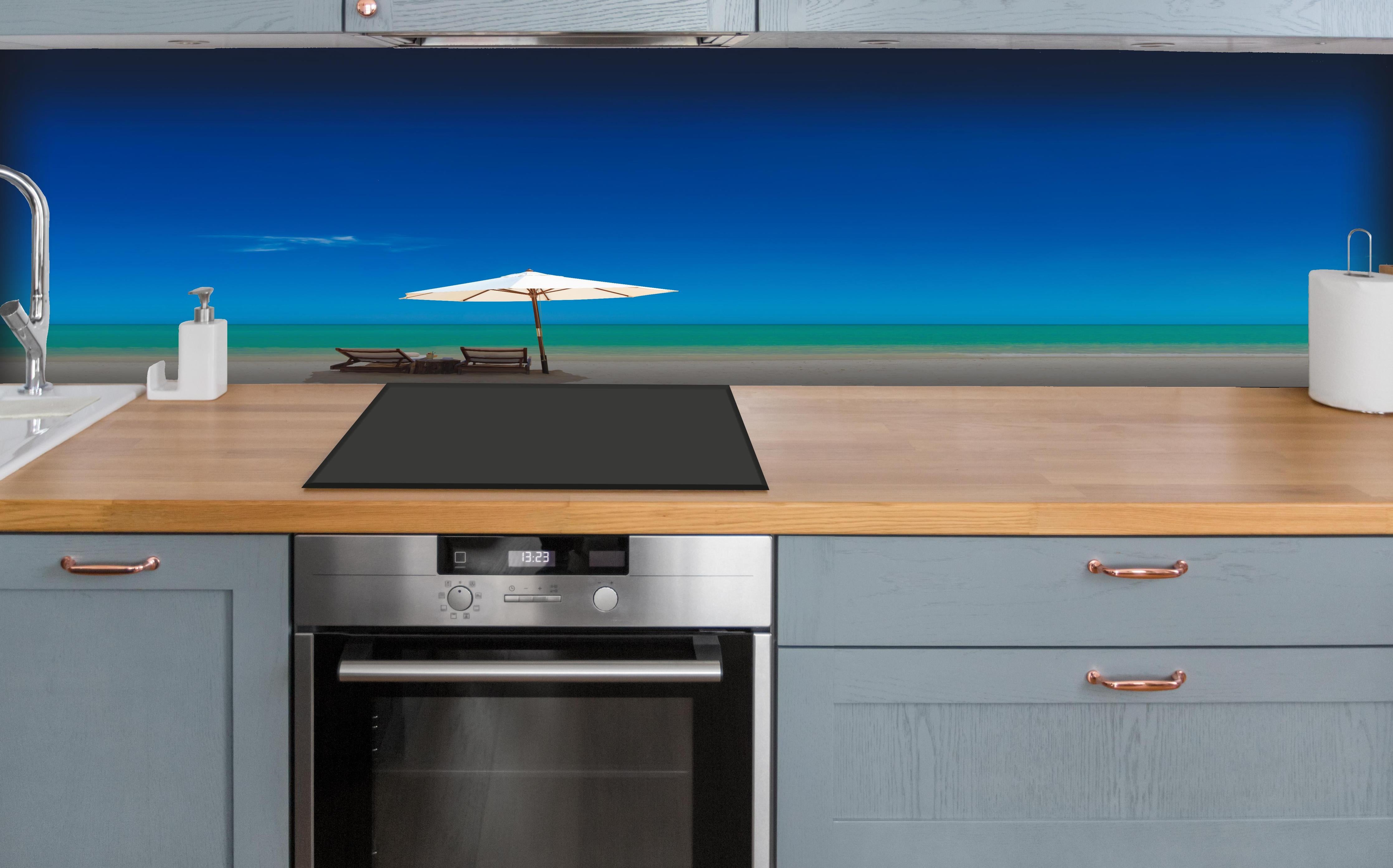 Küche - Idyllischer Strand mit Liegen und Sonnenschirm unter blauem Himmel hinter weißen Hochglanz-Küchenregalen und schwarzem Wasserhahn