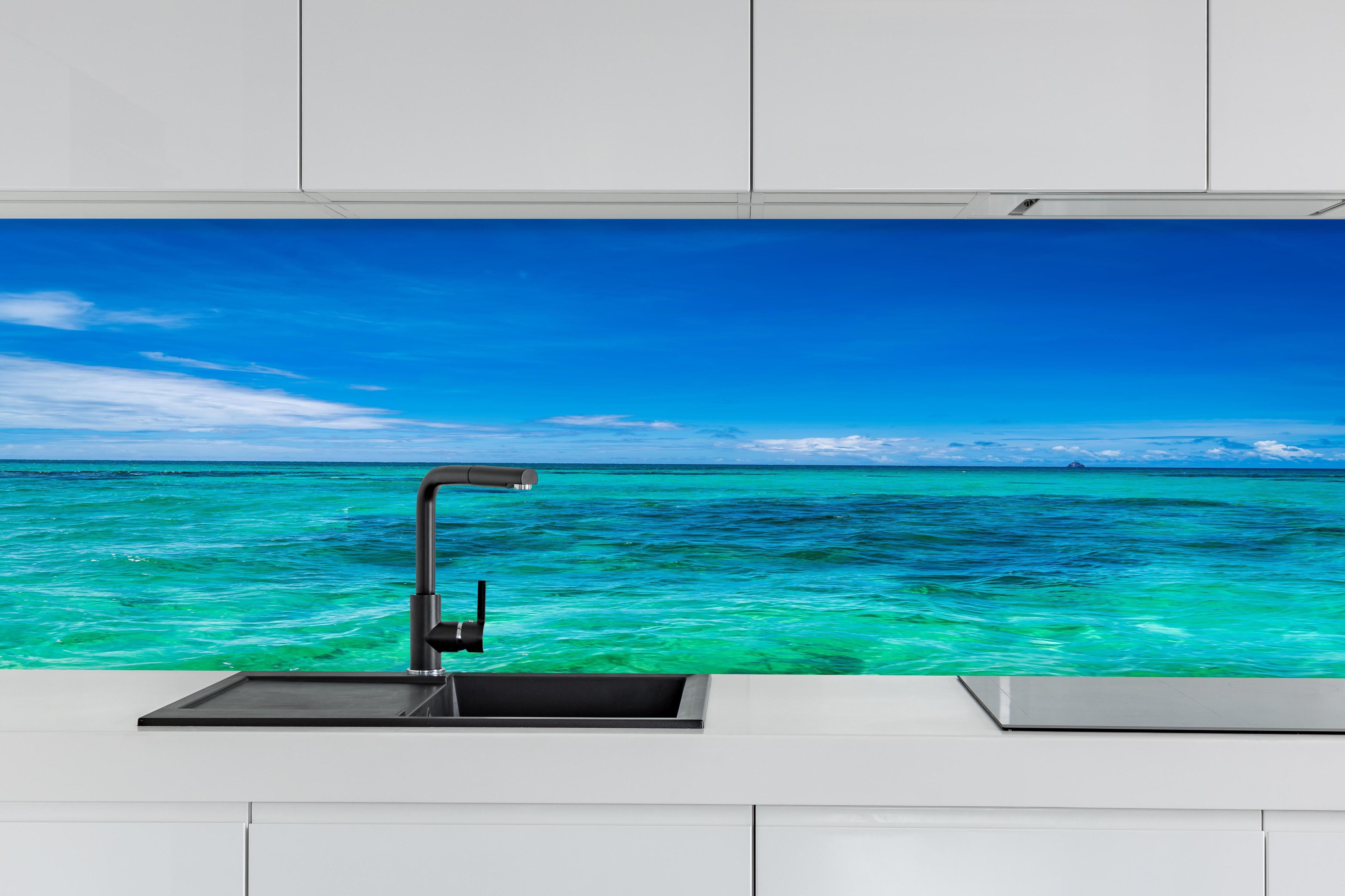 Küche - Klarmarkiertes, türkisblaues Wasser unter hellem Himmel hinter weißen Hochglanz-Küchenregalen und schwarzem Wasserhahn