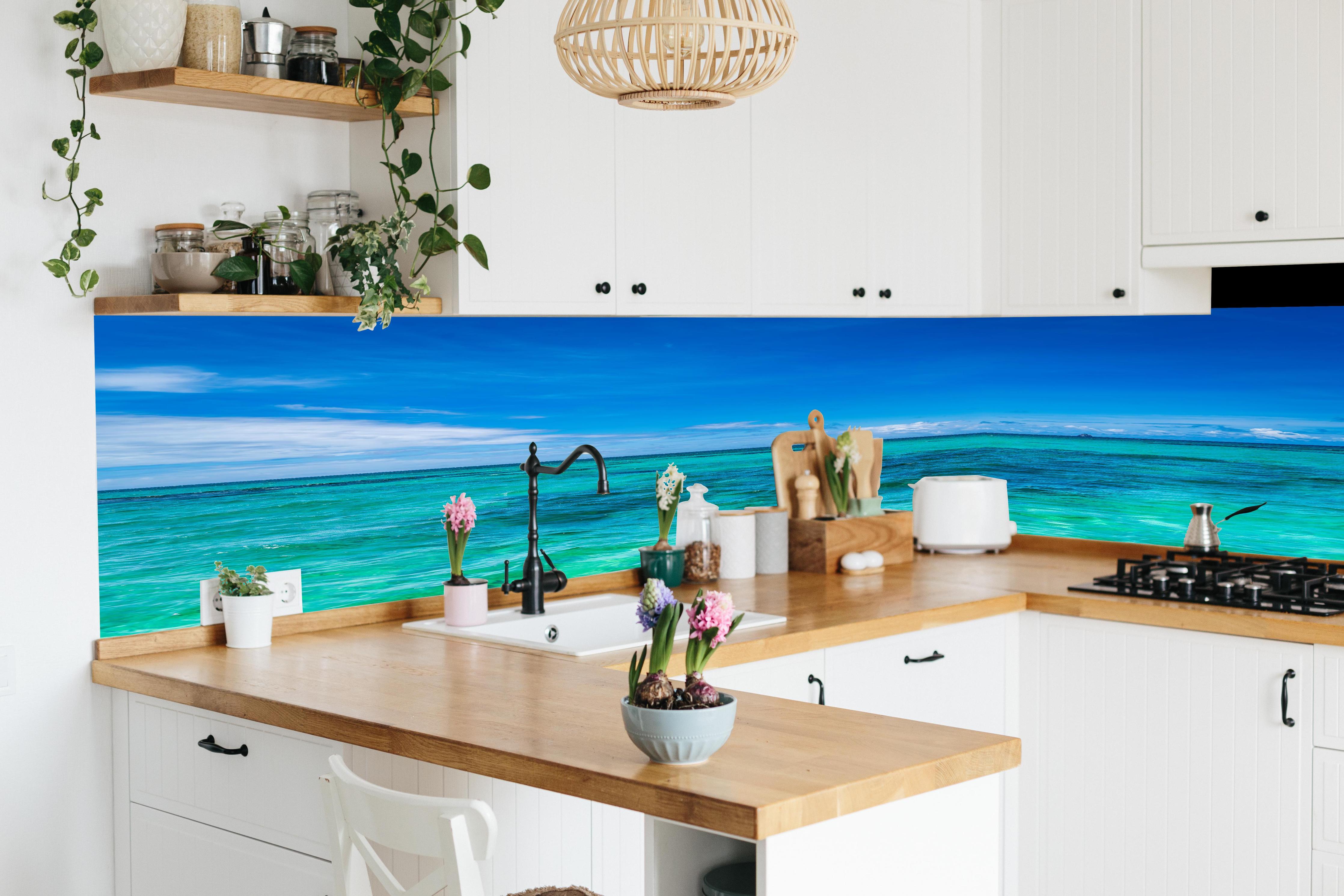 Küche - Klarmarkiertes, türkisblaues Wasser unter hellem Himmel hinter weißen Hochglanz-Küchenregalen und schwarzem Wasserhahn