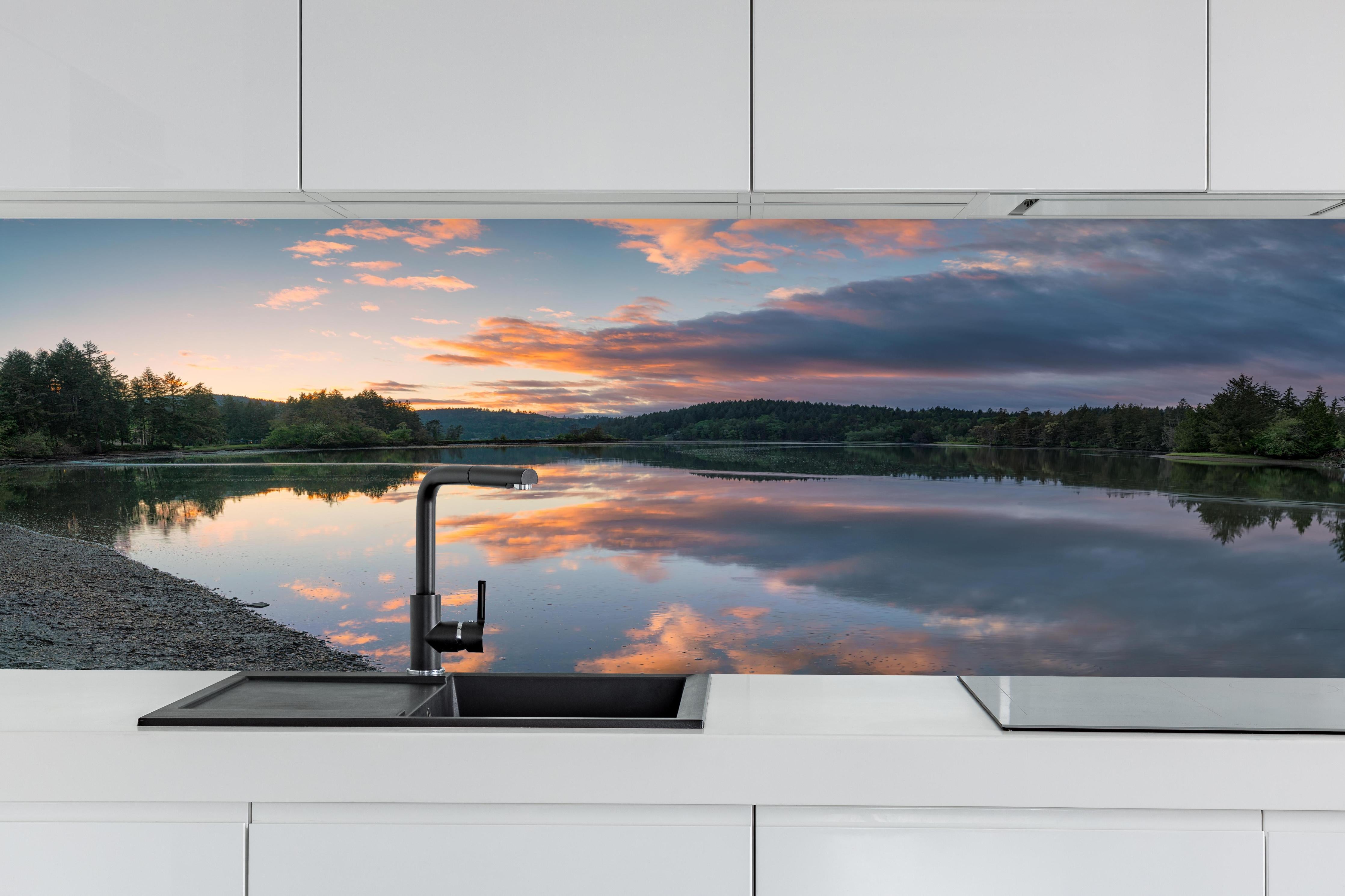 Küche - Spiegelnder See im Sonnenuntergang hinter weißen Hochglanz-Küchenregalen und schwarzem Wasserhahn