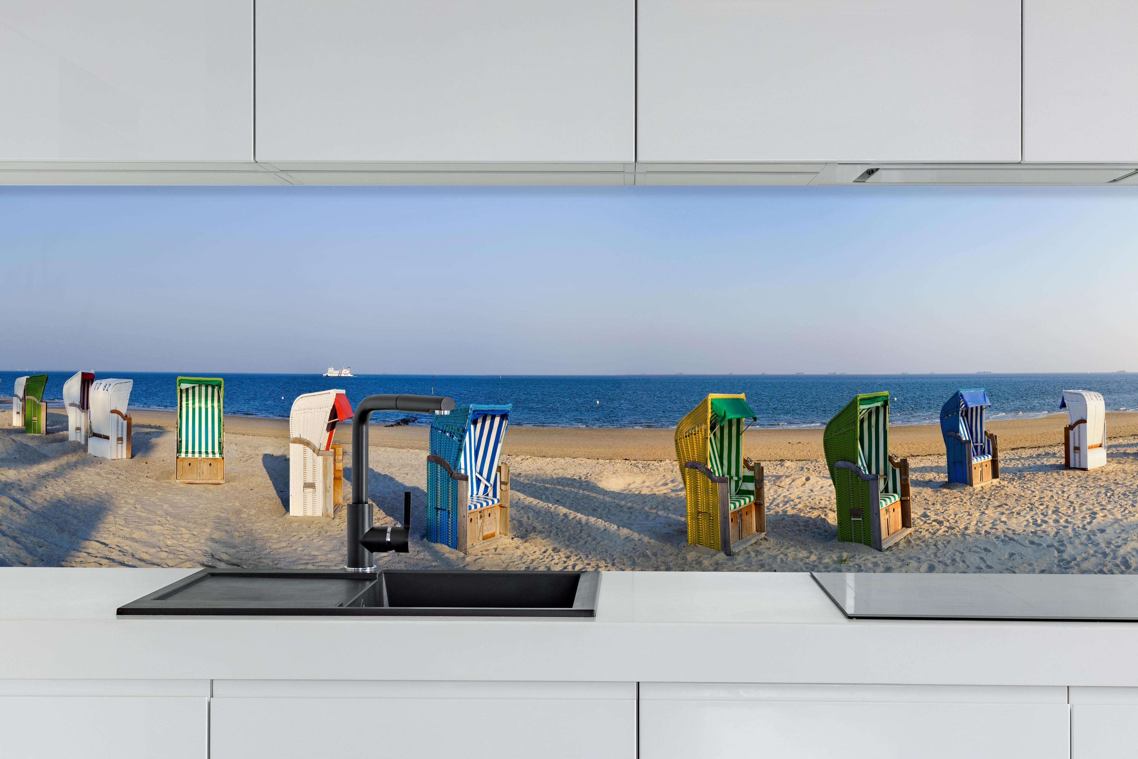 Küche - Kunstvolle, bunte Strandkörbe am Sandstrand mit Blick aufs Meer hinter weißen Hochglanz-Küchenregalen und schwarzem Wasserhahn