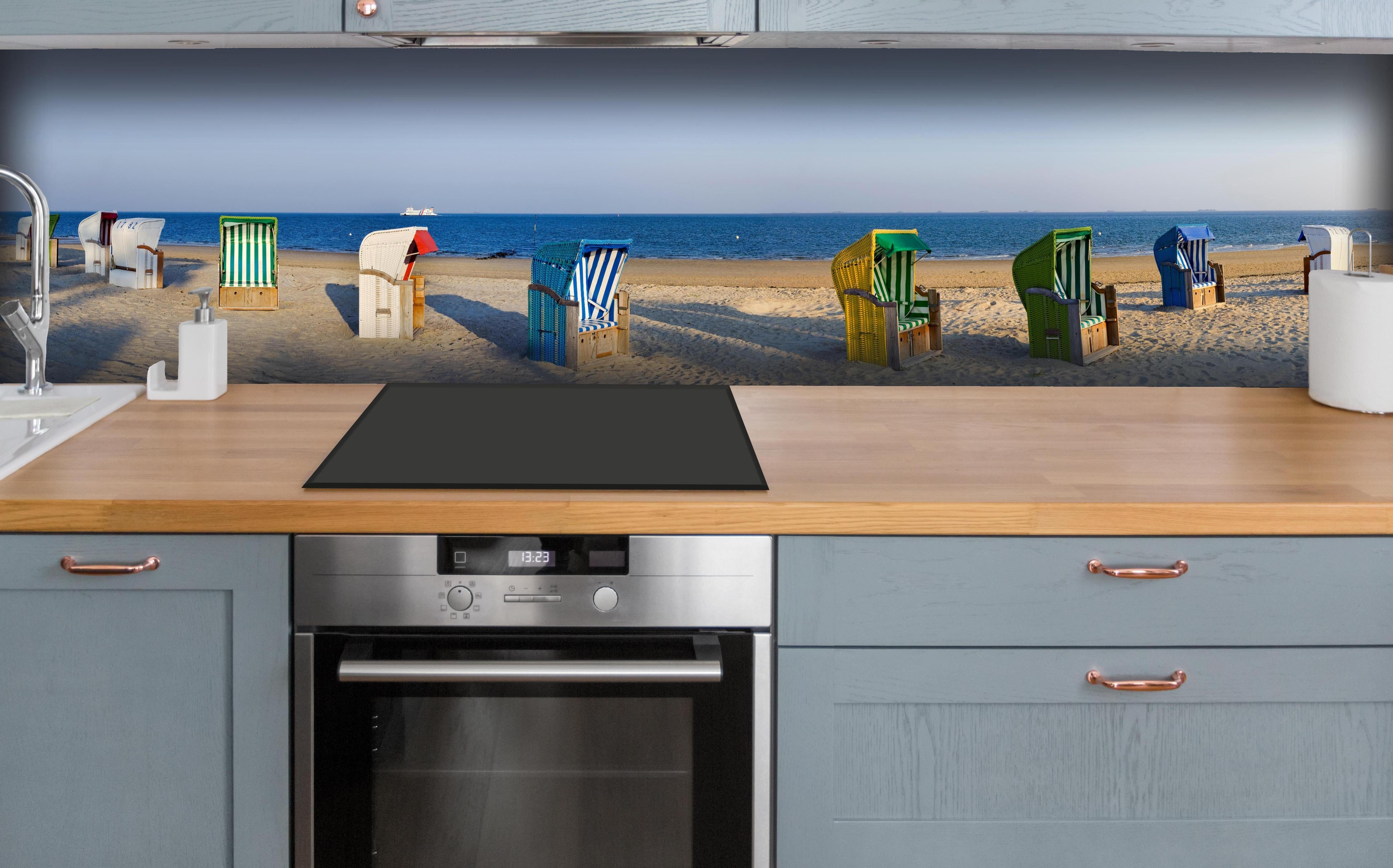 Küche - Kunstvolle, bunte Strandkörbe am Sandstrand mit Blick aufs Meer hinter weißen Hochglanz-Küchenregalen und schwarzem Wasserhahn