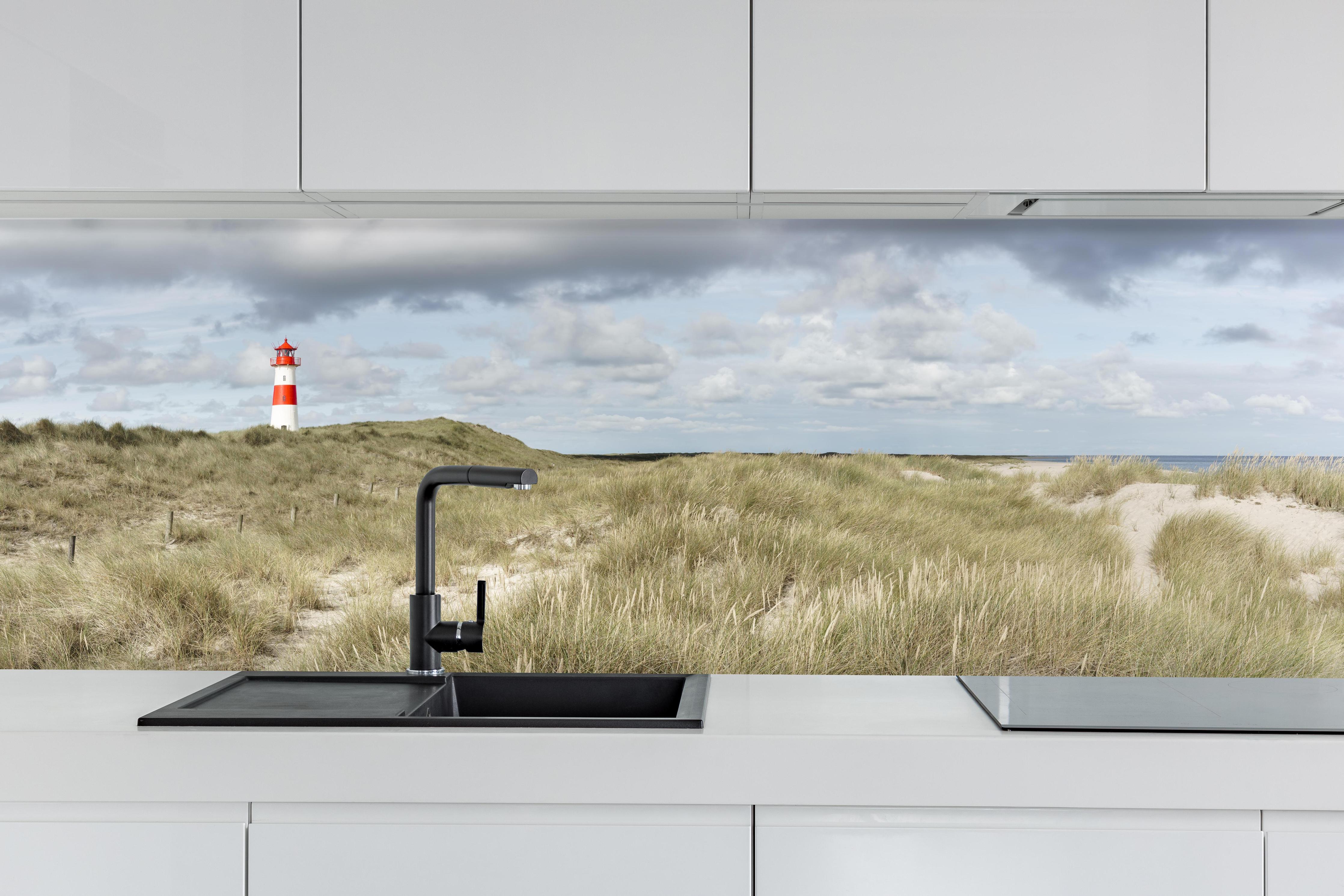 Küche - Malerischer Leuchtturm in den Dünen einer Küstenlandschaft hinter weißen Hochglanz-Küchenregalen und schwarzem Wasserhahn
