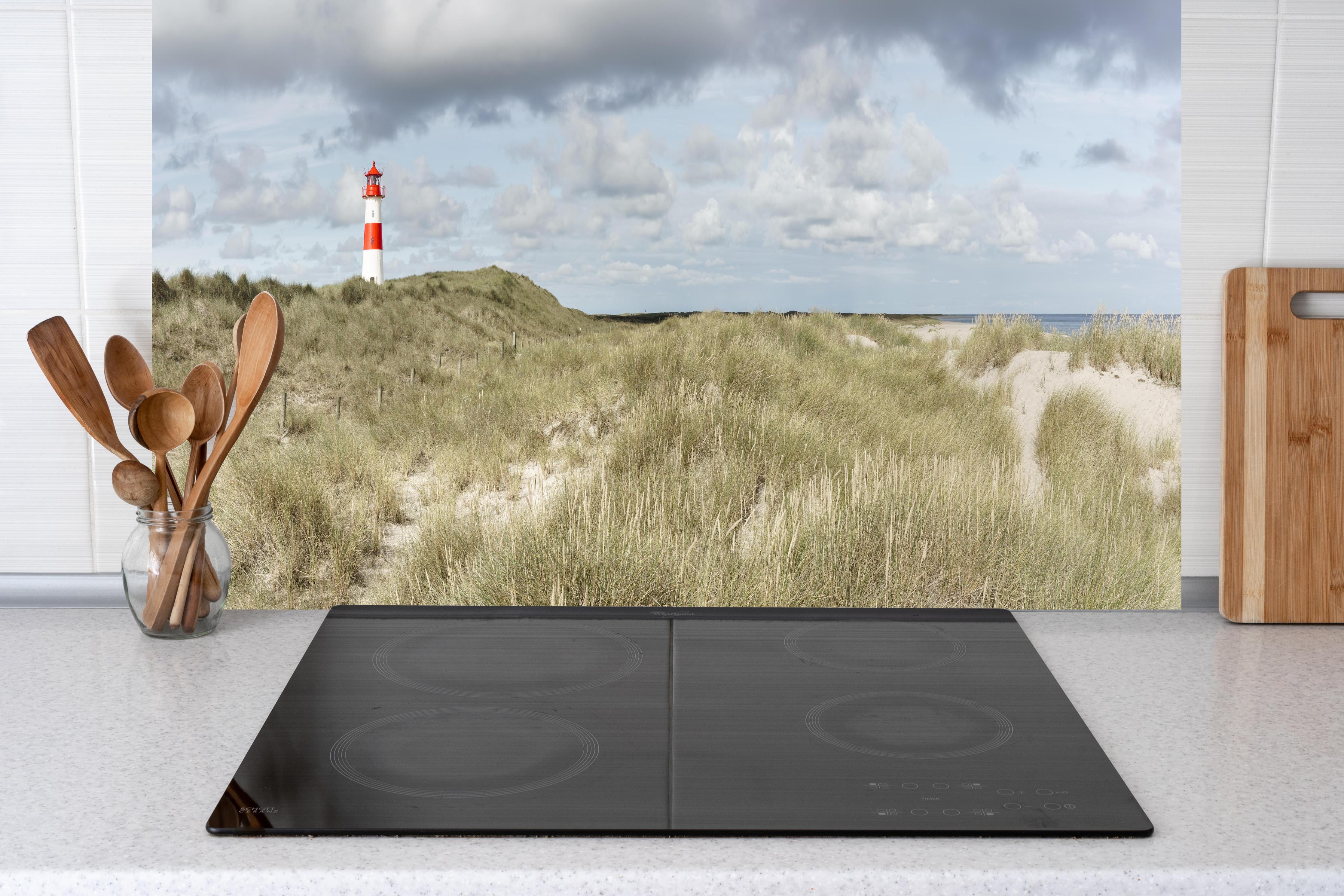 Küche - Malerischer Leuchtturm in den Dünen einer Küstenlandschaft hinter weißen Hochglanz-Küchenregalen und schwarzem Wasserhahn