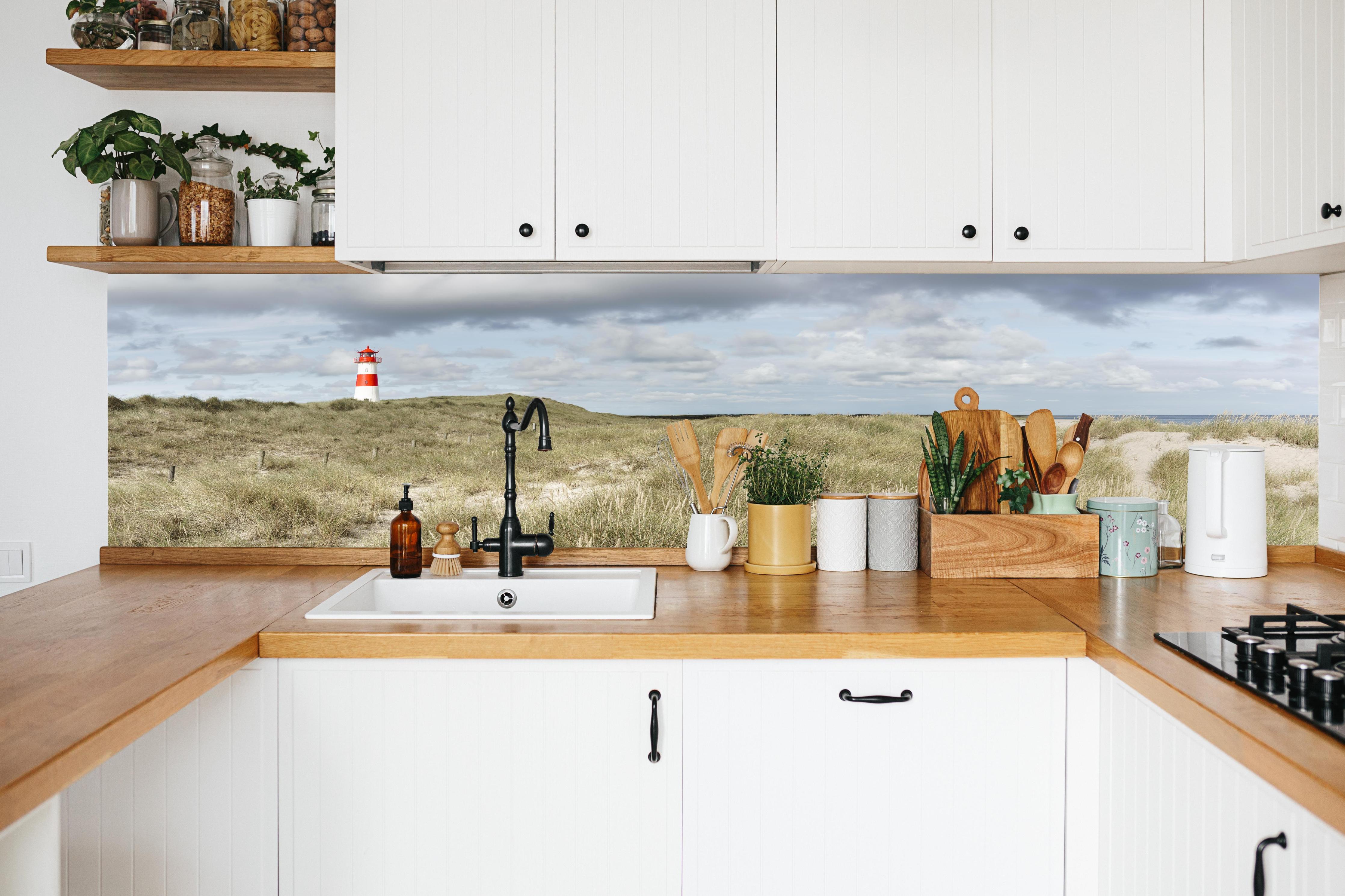 Küche - Malerischer Leuchtturm in den Dünen einer Küstenlandschaft hinter weißen Hochglanz-Küchenregalen und schwarzem Wasserhahn