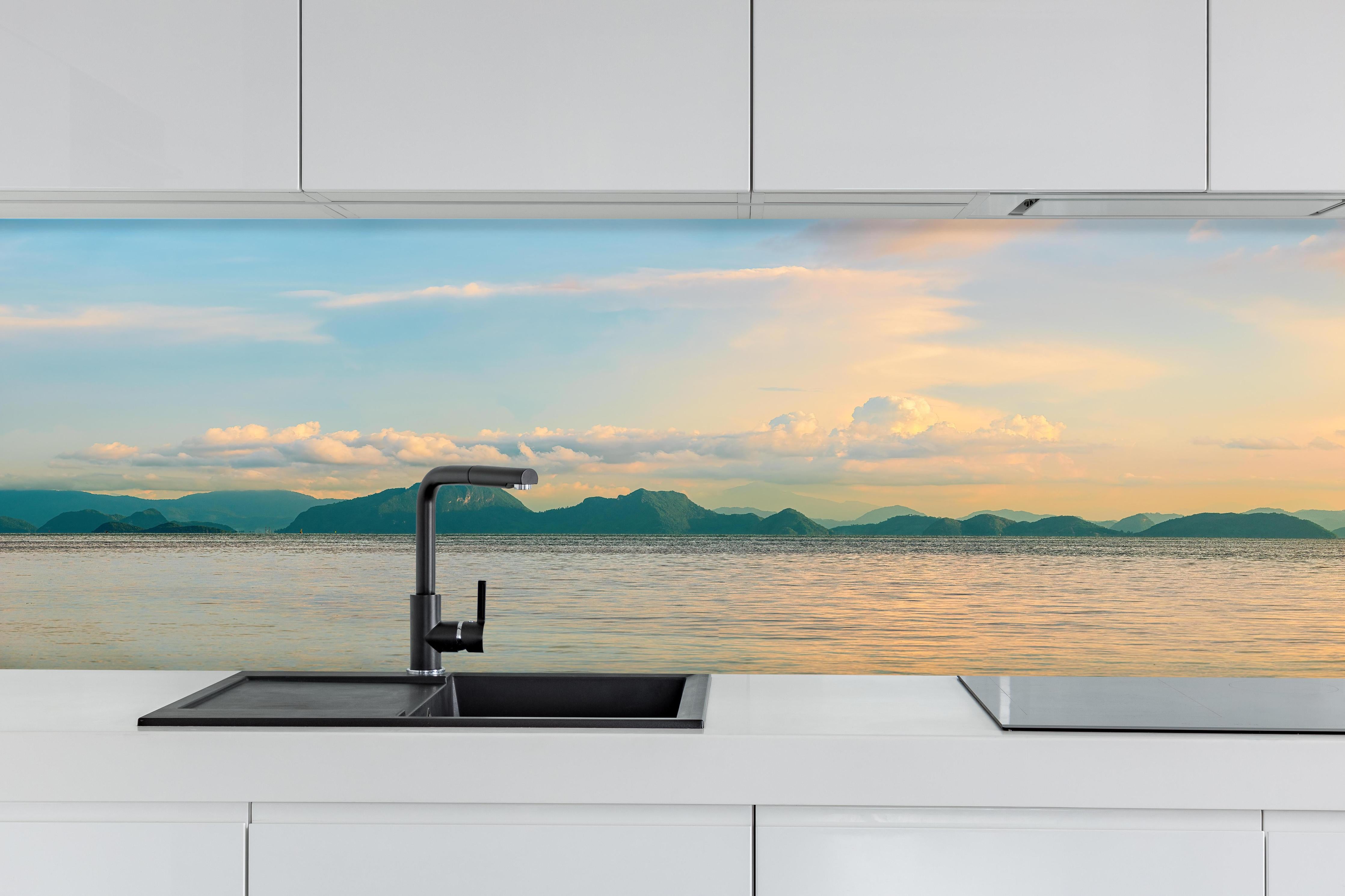 Küche - Panoramablick auf idyllische Küstenlandschaft mit Berge im Hintergrund hinter weißen Hochglanz-Küchenregalen und schwarzem Wasserhahn
