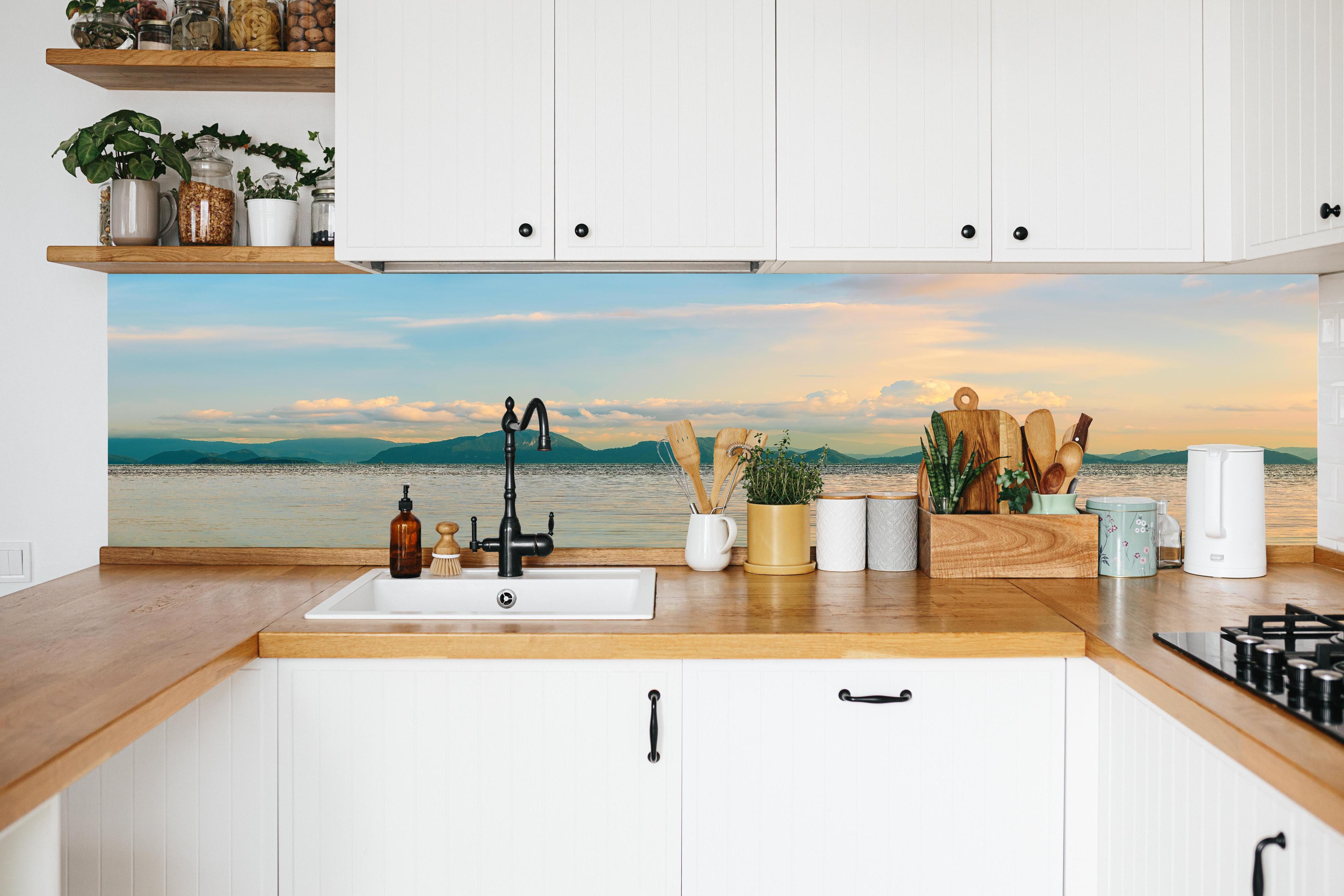Küche - Panoramablick auf idyllische Küstenlandschaft mit Berge im Hintergrund hinter weißen Hochglanz-Küchenregalen und schwarzem Wasserhahn