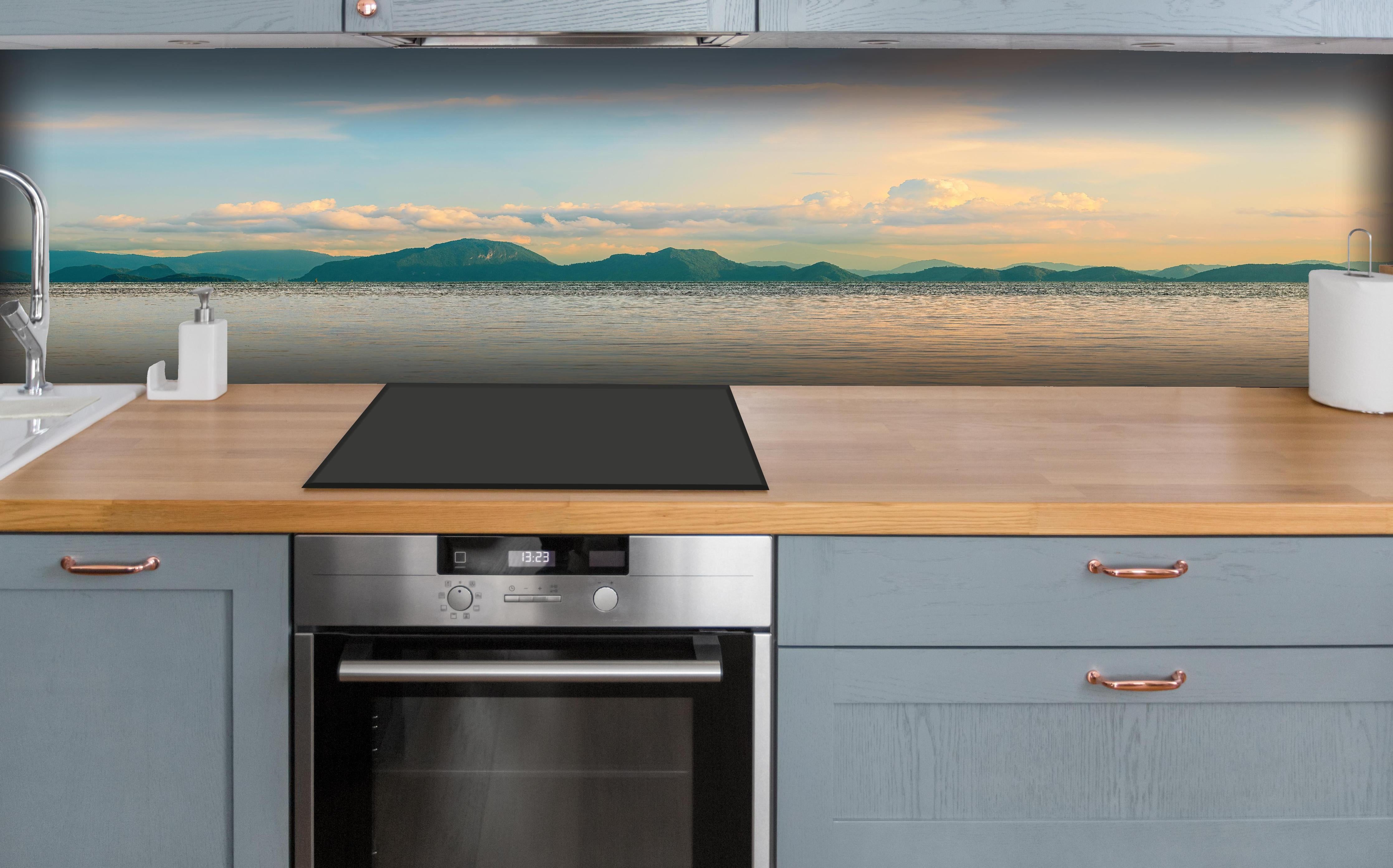 Küche - Panoramablick auf idyllische Küstenlandschaft mit Berge im Hintergrund hinter weißen Hochglanz-Küchenregalen und schwarzem Wasserhahn