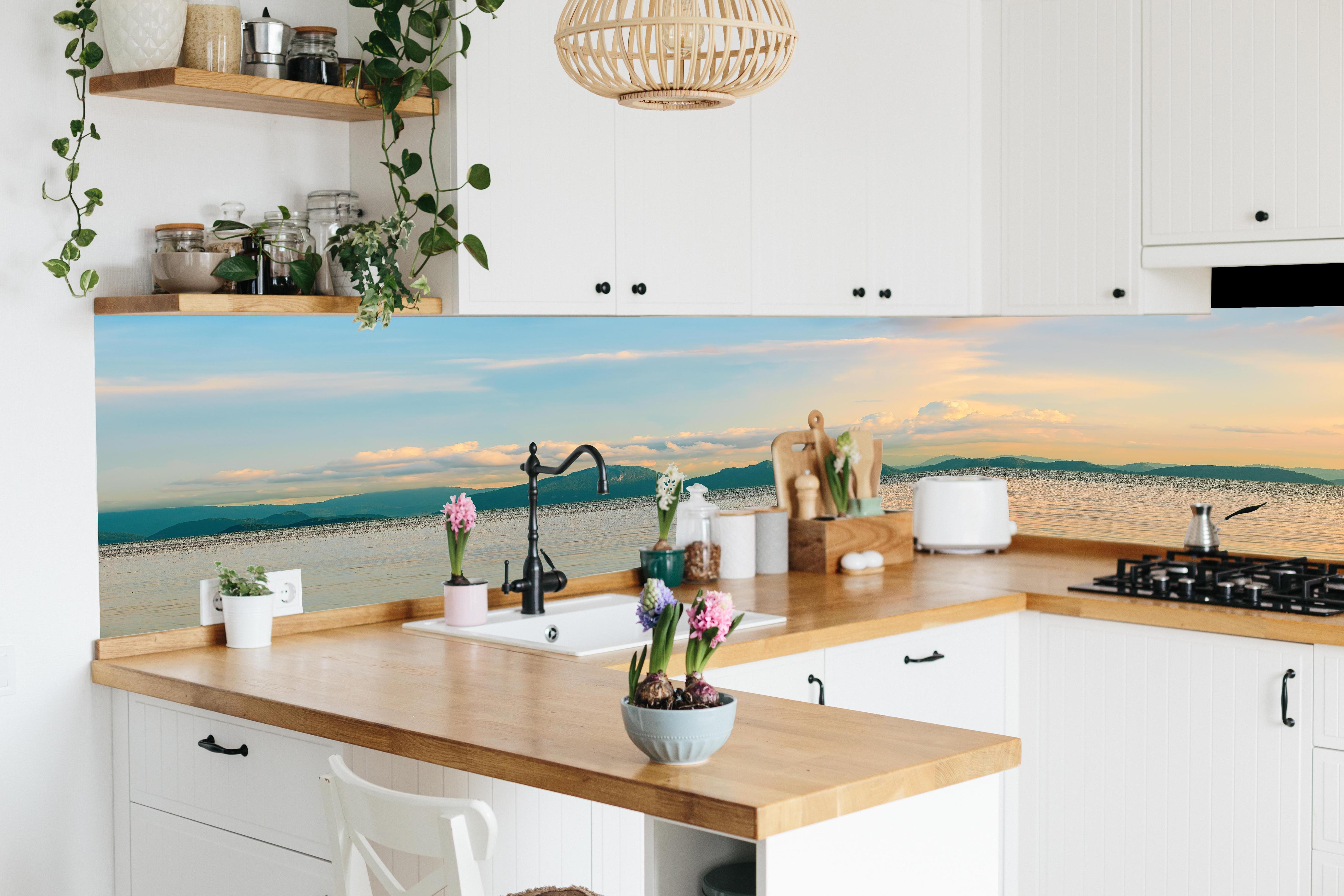 Küche - Panoramablick auf idyllische Küstenlandschaft mit Berge im Hintergrund hinter weißen Hochglanz-Küchenregalen und schwarzem Wasserhahn