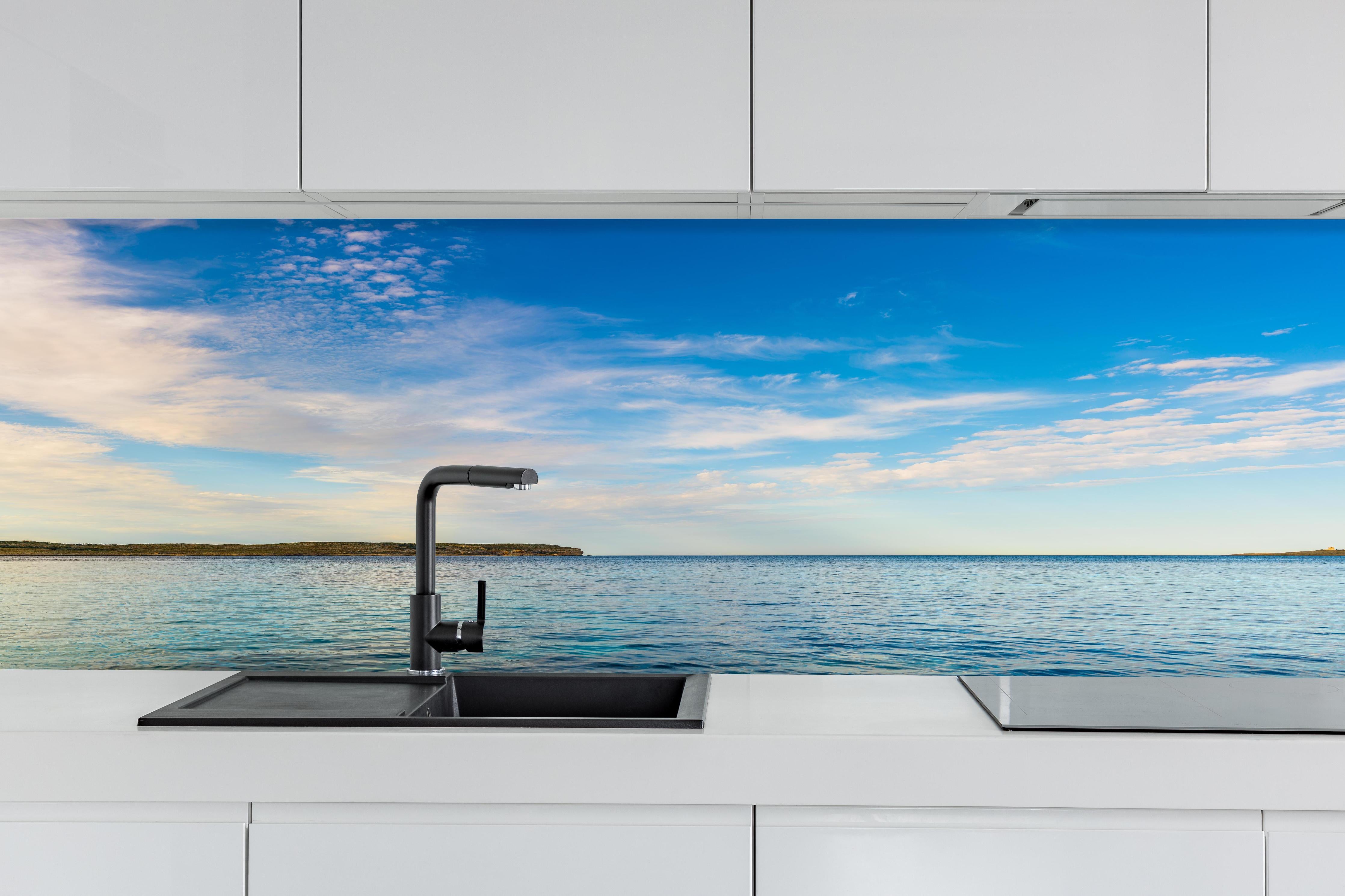 Küche - Weite und ruhige Meerelandschaft unter einem strahlend blauen Himmel hinter weißen Hochglanz-Küchenregalen und schwarzem Wasserhahn