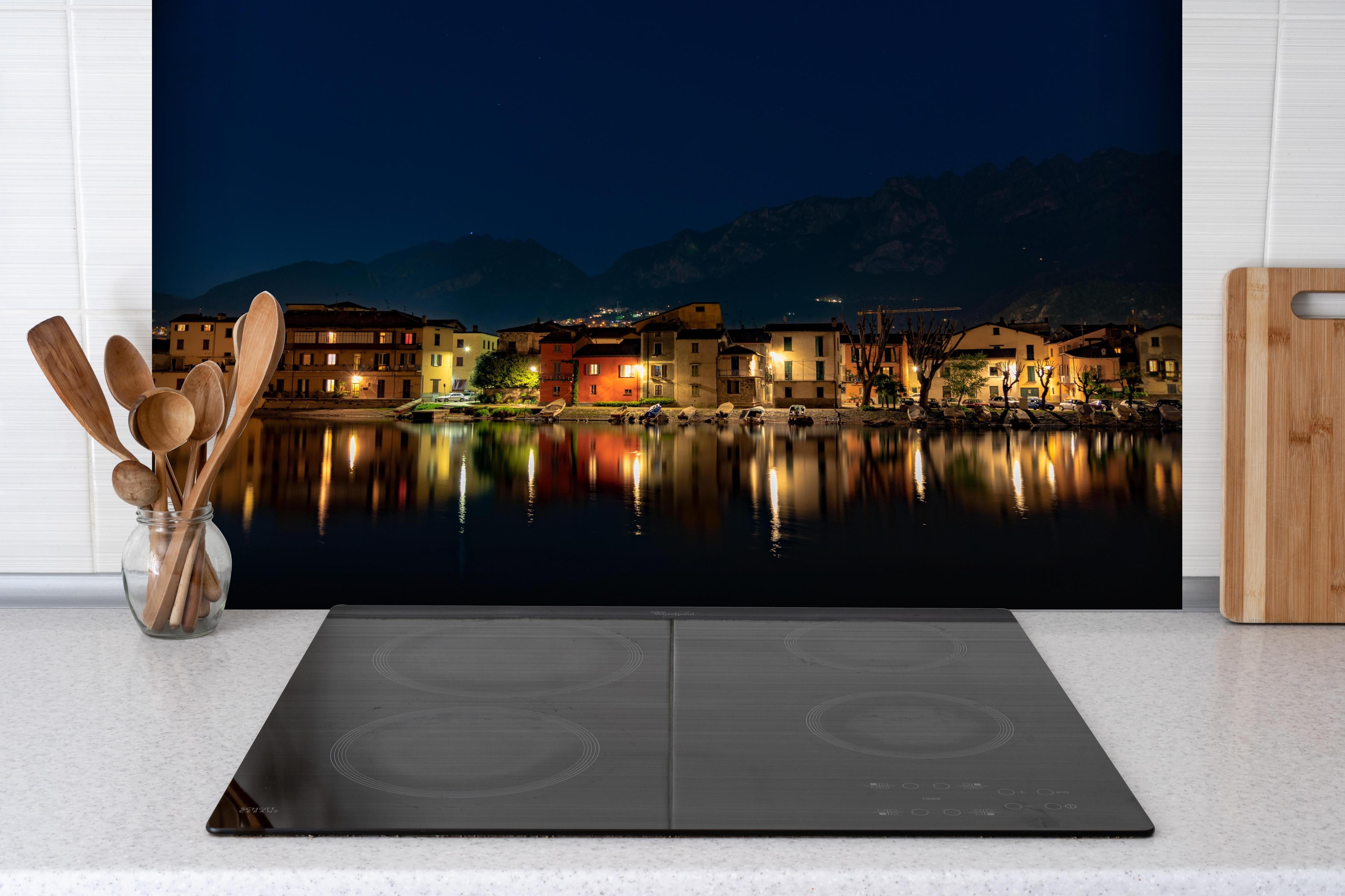 Spritzschutz - Beleuchtete Seelandschaft bei Nacht hinter einem Cerankochfeld zwischen Holz-Kochutensilien