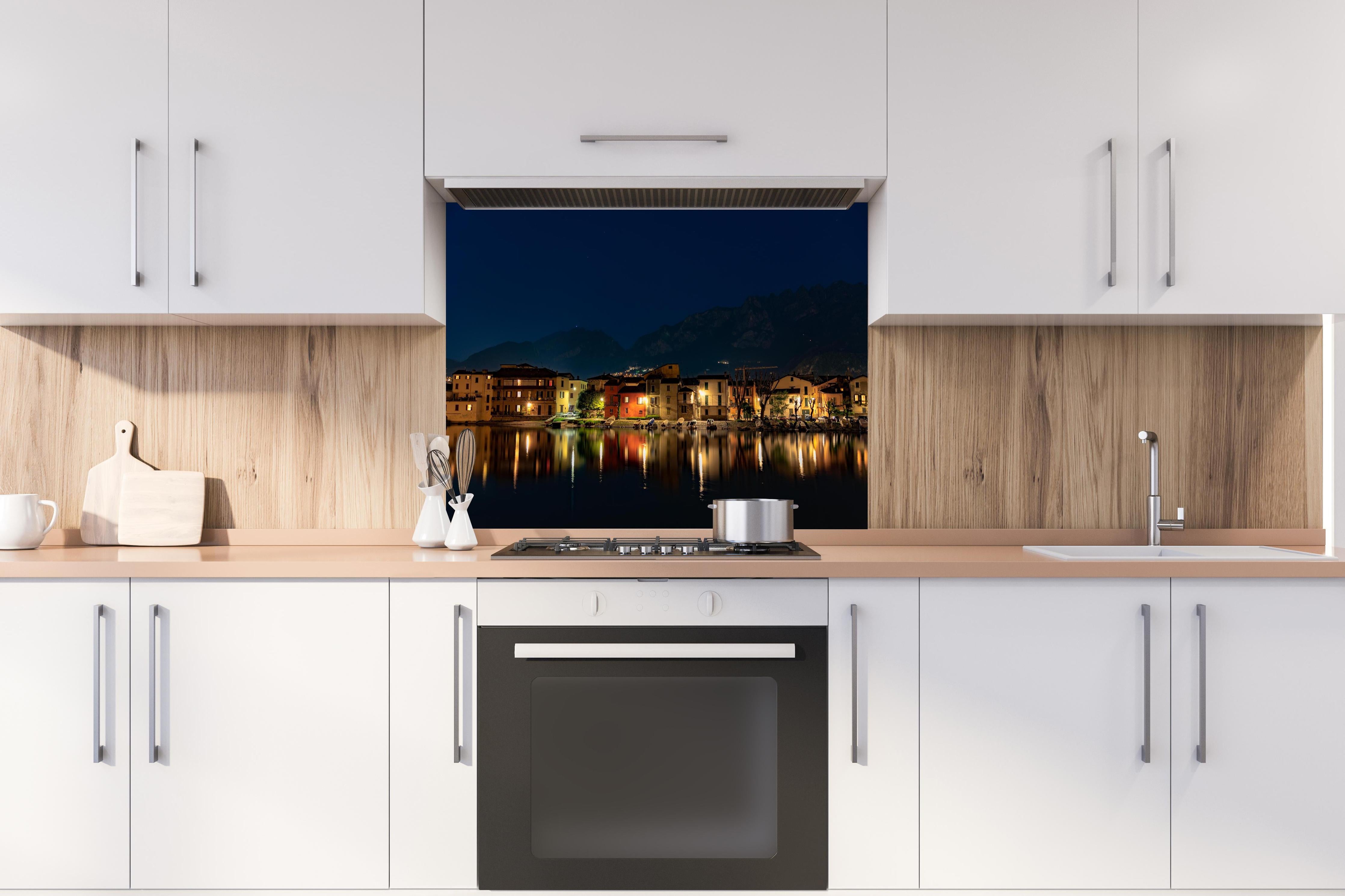 Spritzschutz - Beleuchtete Seelandschaft bei Nacht hinter einem Cerankochfeld zwischen Holz-Kochutensilien