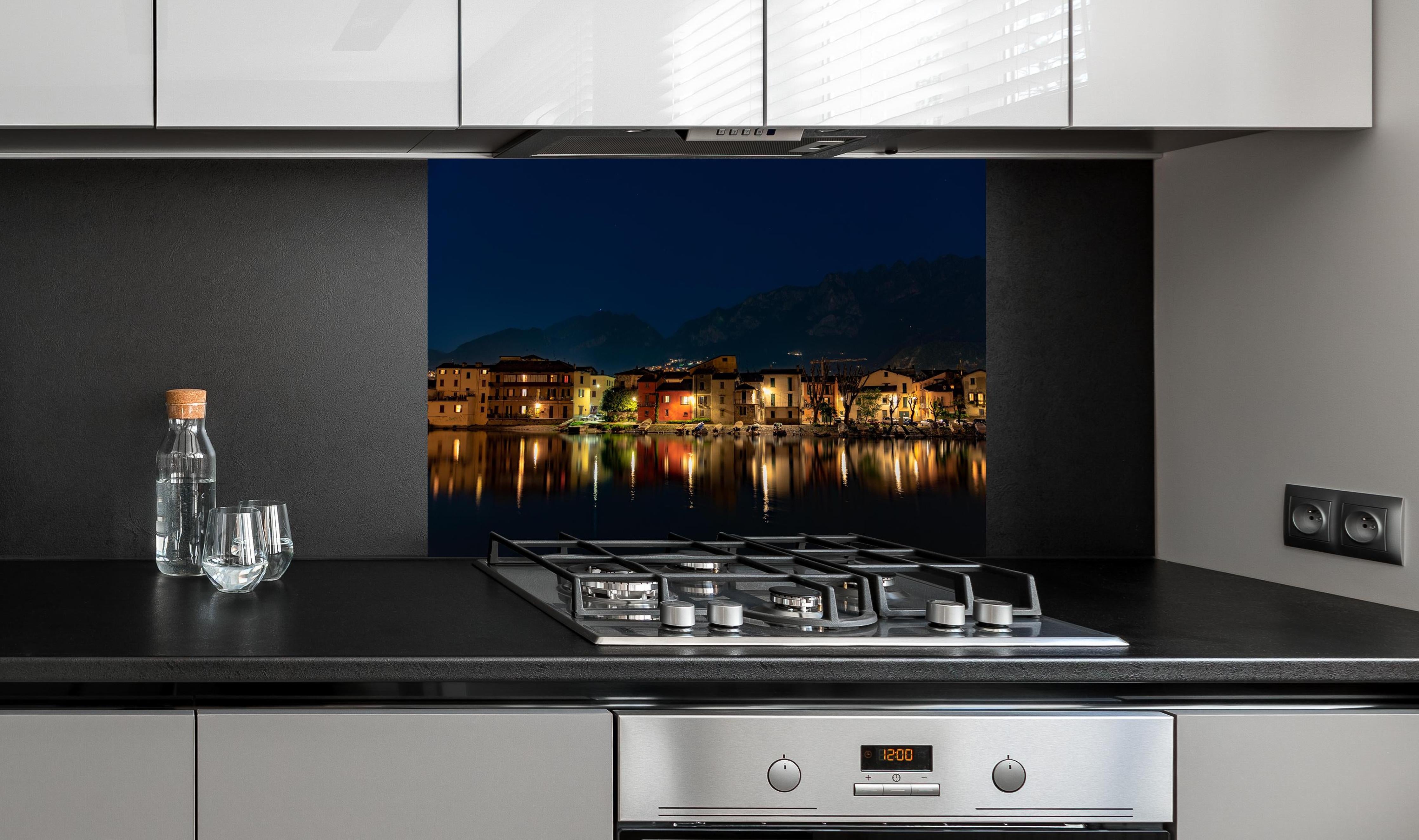 Spritzschutz - Beleuchtete Seelandschaft bei Nacht hinter einem Cerankochfeld zwischen Holz-Kochutensilien