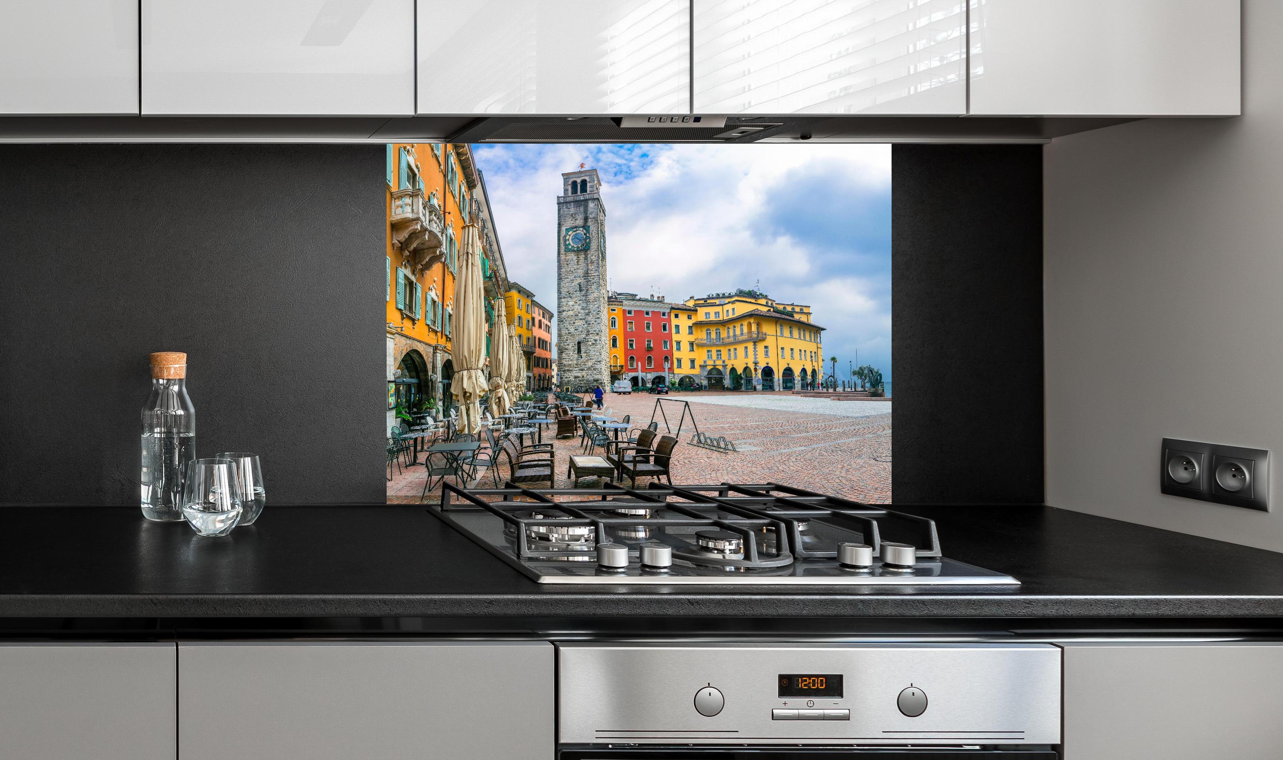 Spritzschutz - Charmante Stadt am Gardasee - Riva del Garda hinter einem Cerankochfeld zwischen Holz-Kochutensilien