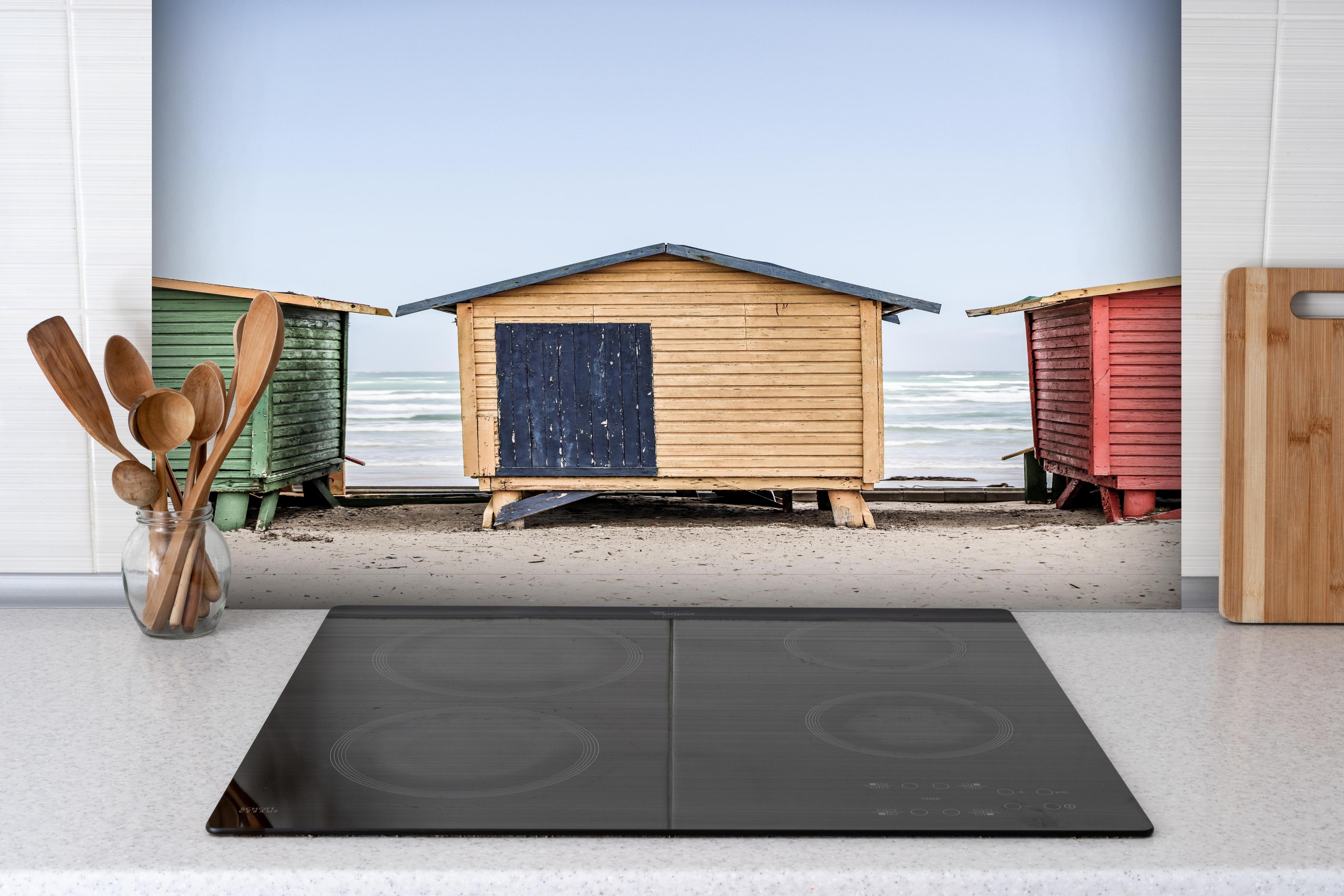 Spritzschutz - Farbenfrohe Strandhütten am Meeressand hinter einem Cerankochfeld zwischen Holz-Kochutensilien