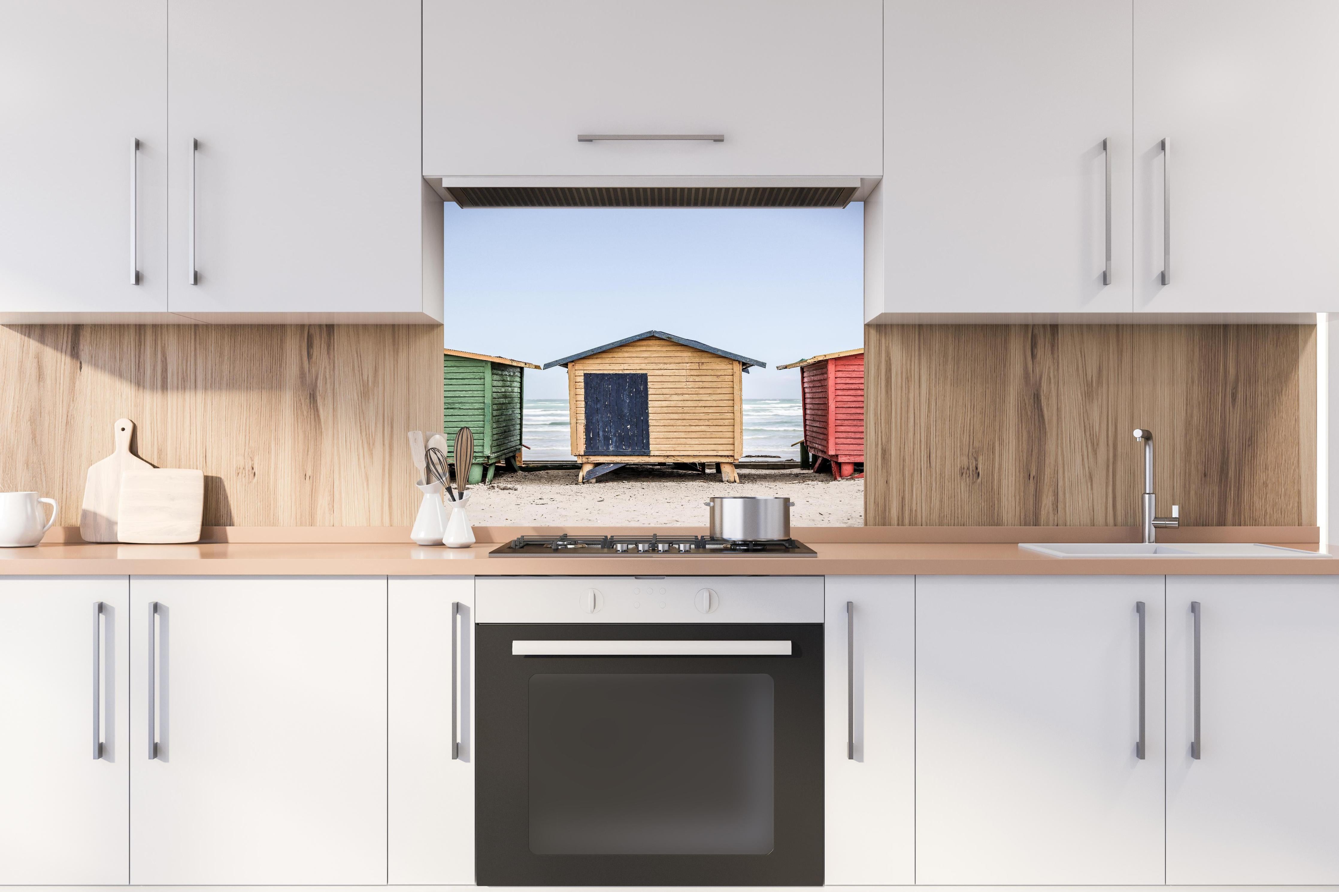 Spritzschutz - Farbenfrohe Strandhütten am Meeressand hinter einem Cerankochfeld zwischen Holz-Kochutensilien