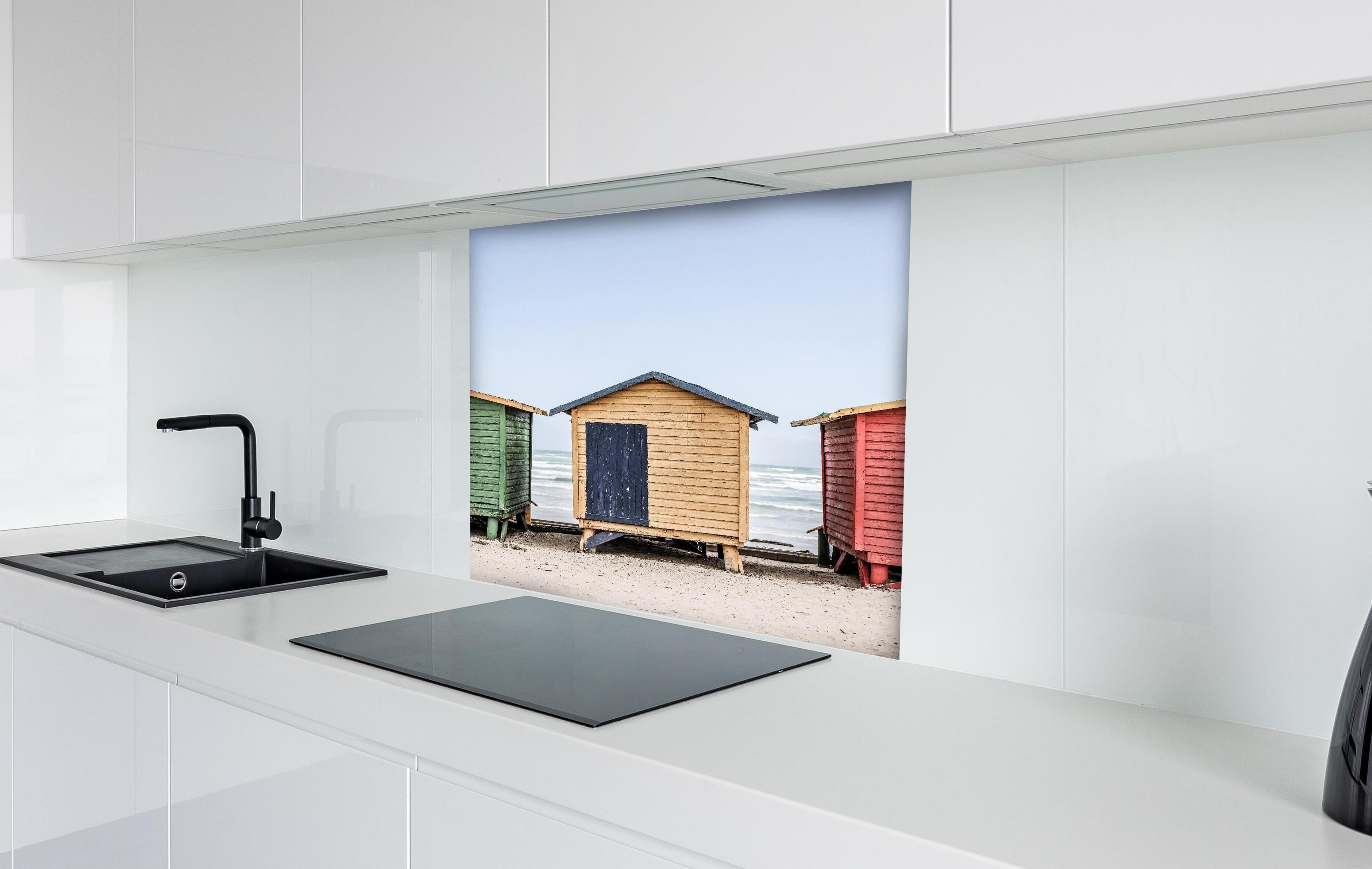 Spritzschutz - Farbenfrohe Strandhütten am Meeressand hinter einem Cerankochfeld zwischen Holz-Kochutensilien