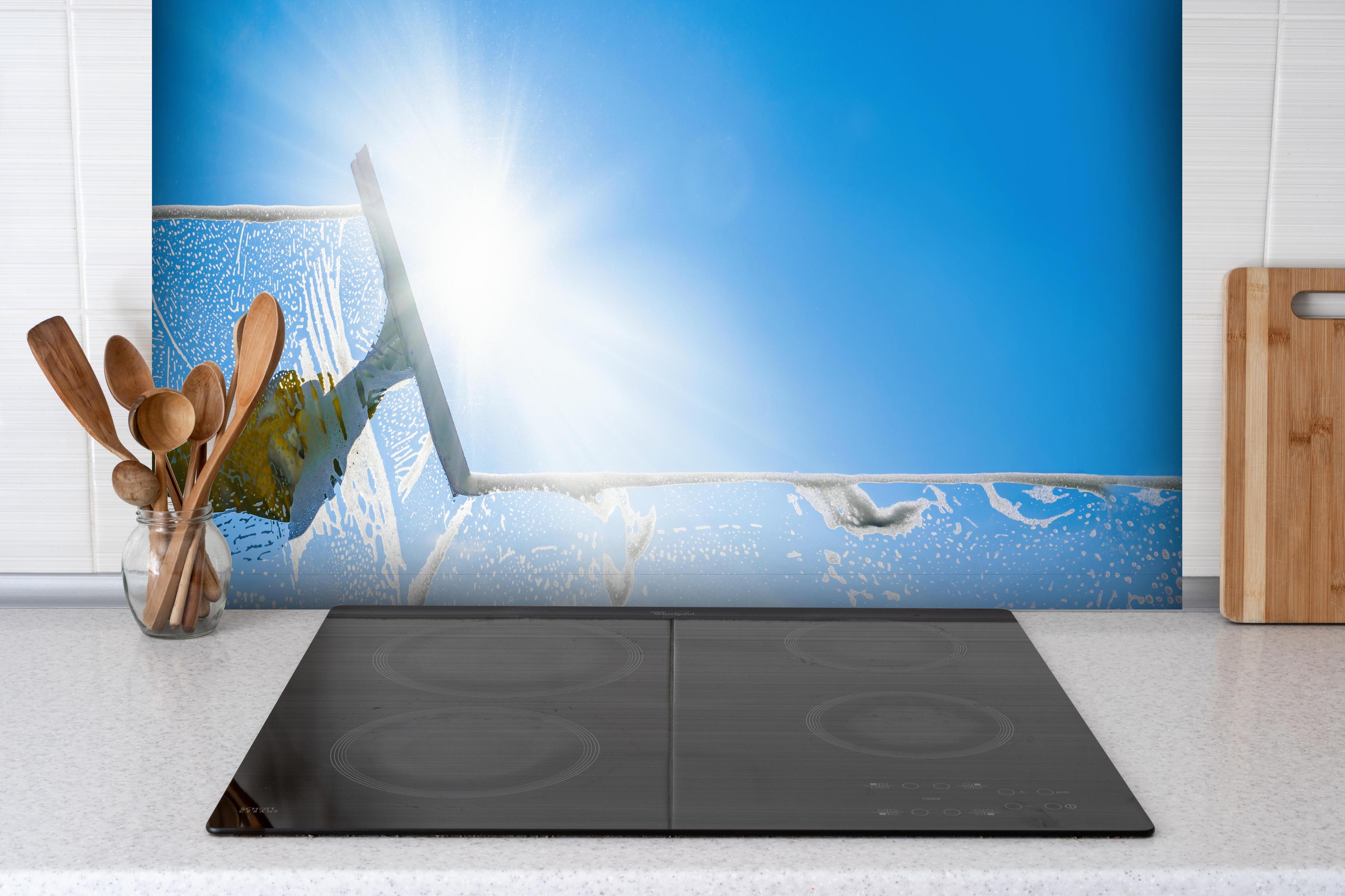 Spritzschutz - Fensterputzer mit Abzieher und strahlendem Himmel hinter einem Cerankochfeld zwischen Holz-Kochutensilien