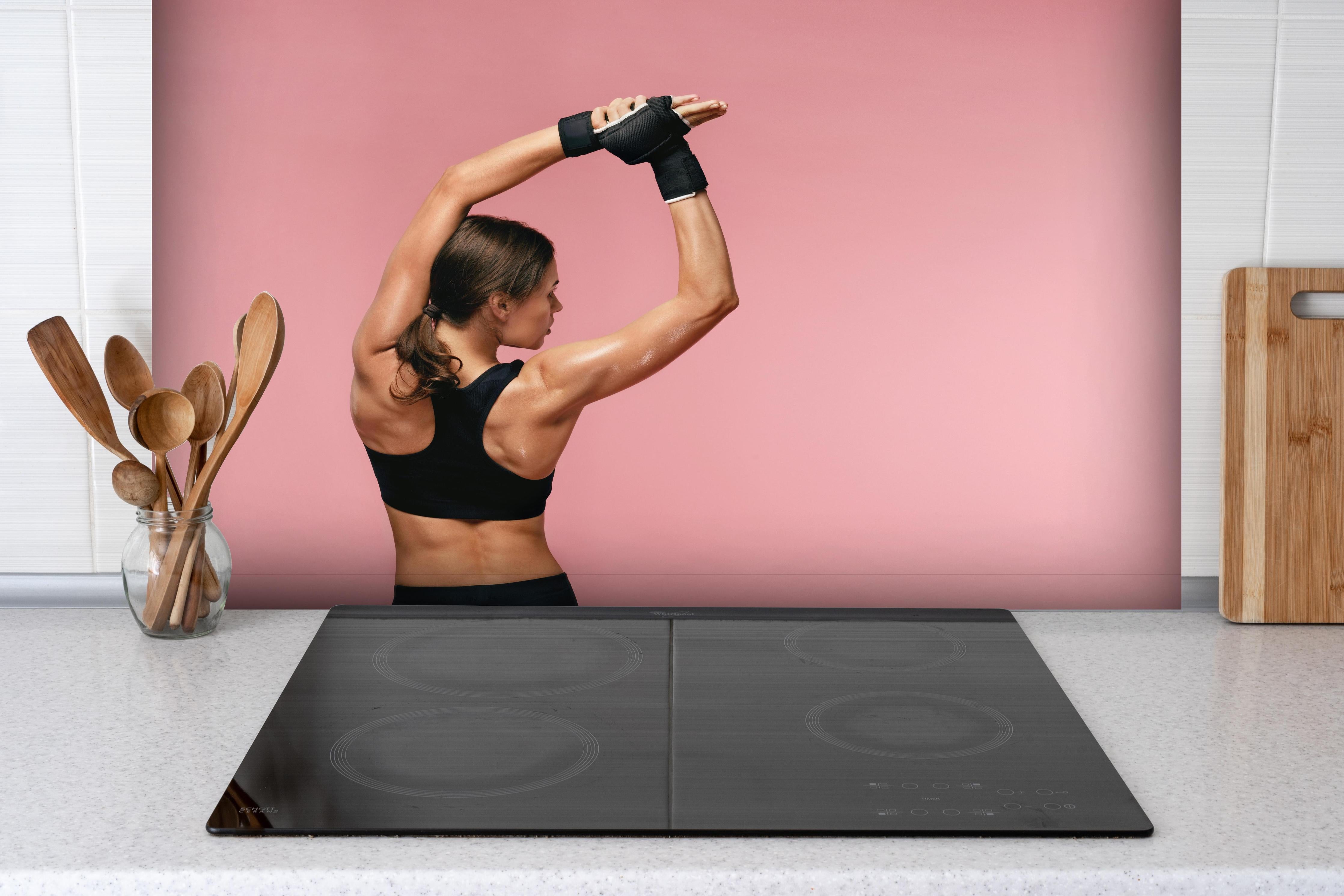 Spritzschutz - Fitness Frau streckt sich vor Training hinter einem Cerankochfeld zwischen Holz-Kochutensilien