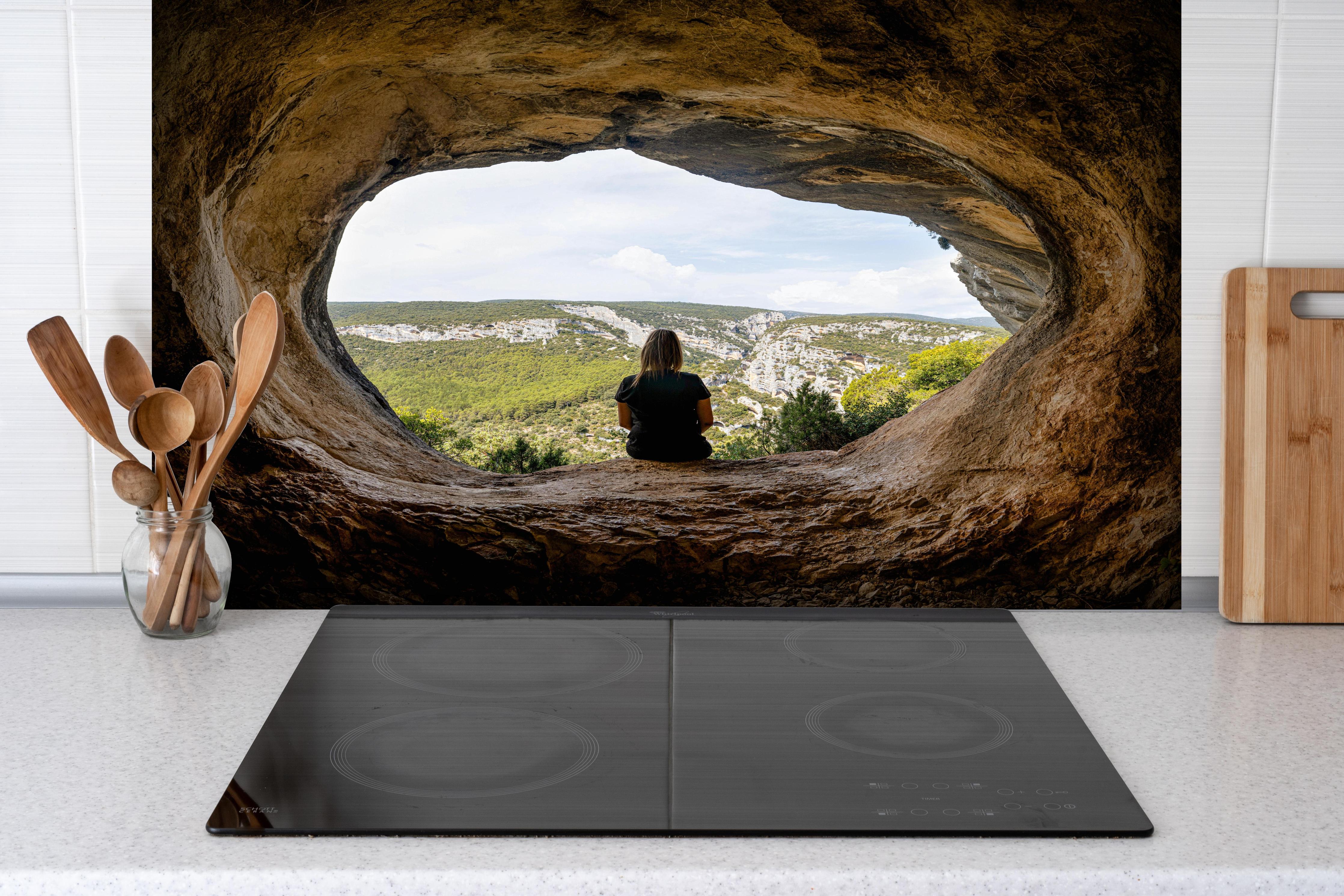 Spritzschutz - Frau sitzend in Berghöhle mit Blick auf Landschaft hinter einem Cerankochfeld zwischen Holz-Kochutensilien
