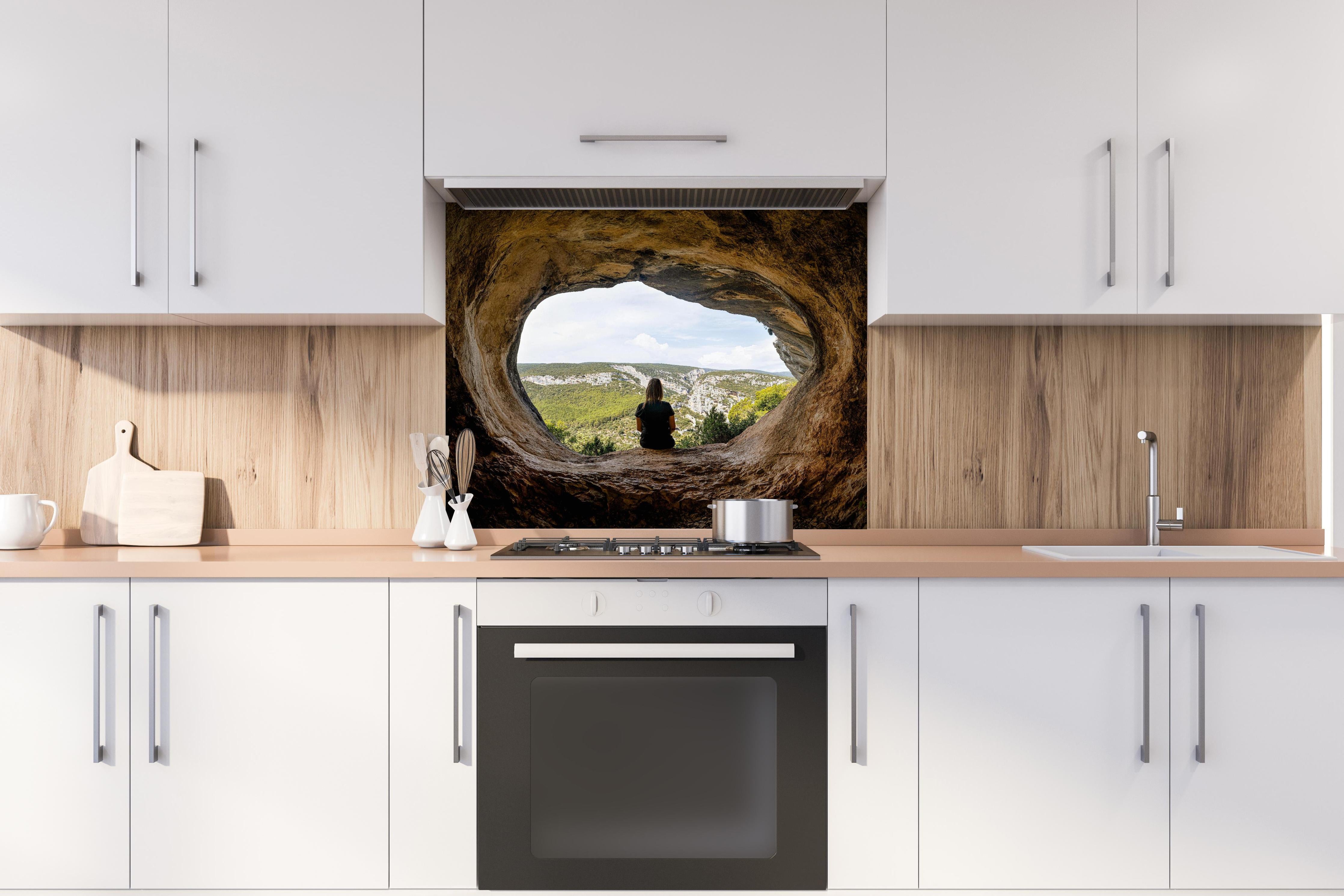 Spritzschutz - Frau sitzend in Berghöhle mit Blick auf Landschaft hinter einem Cerankochfeld zwischen Holz-Kochutensilien