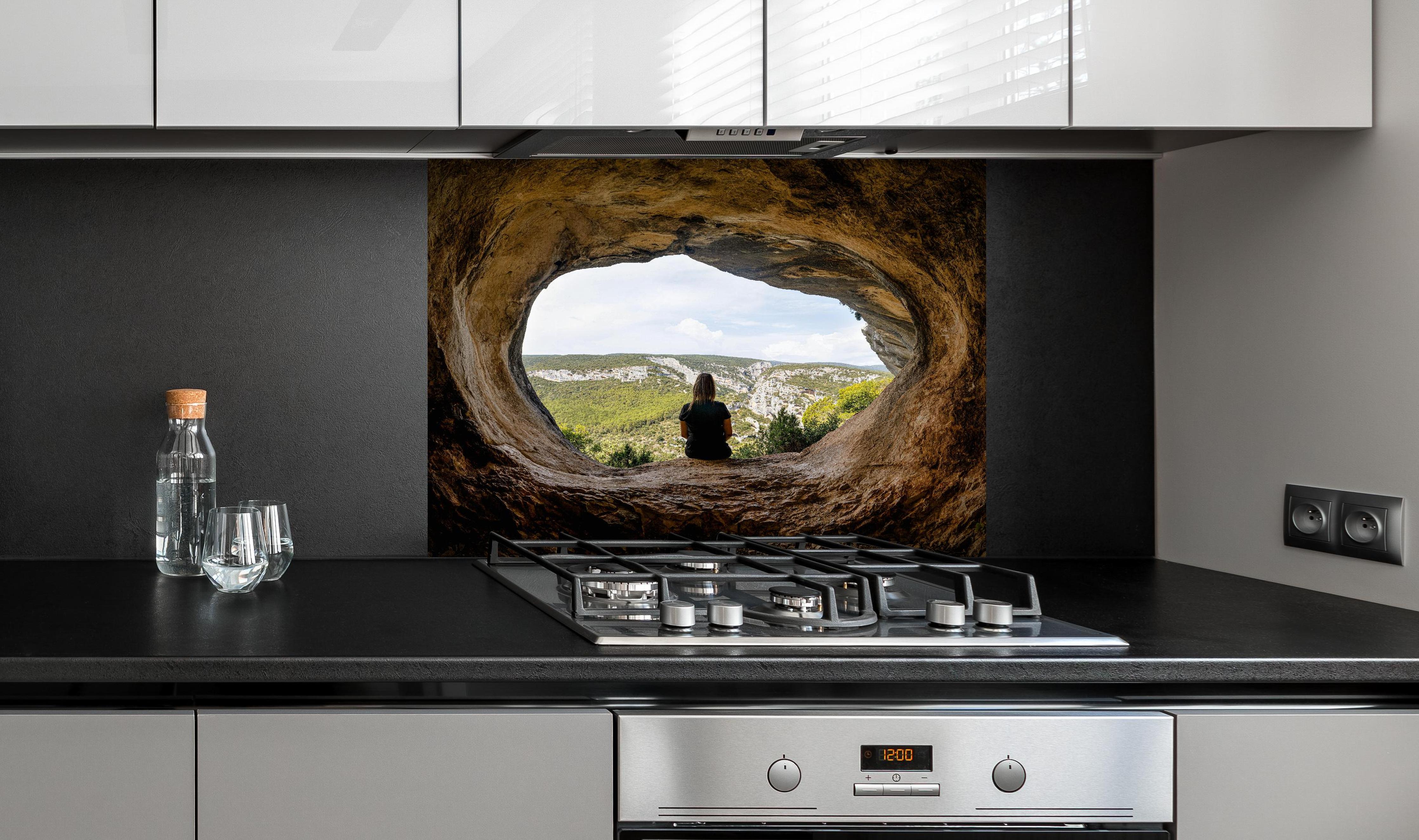 Spritzschutz - Frau sitzend in Berghöhle mit Blick auf Landschaft hinter einem Cerankochfeld zwischen Holz-Kochutensilien