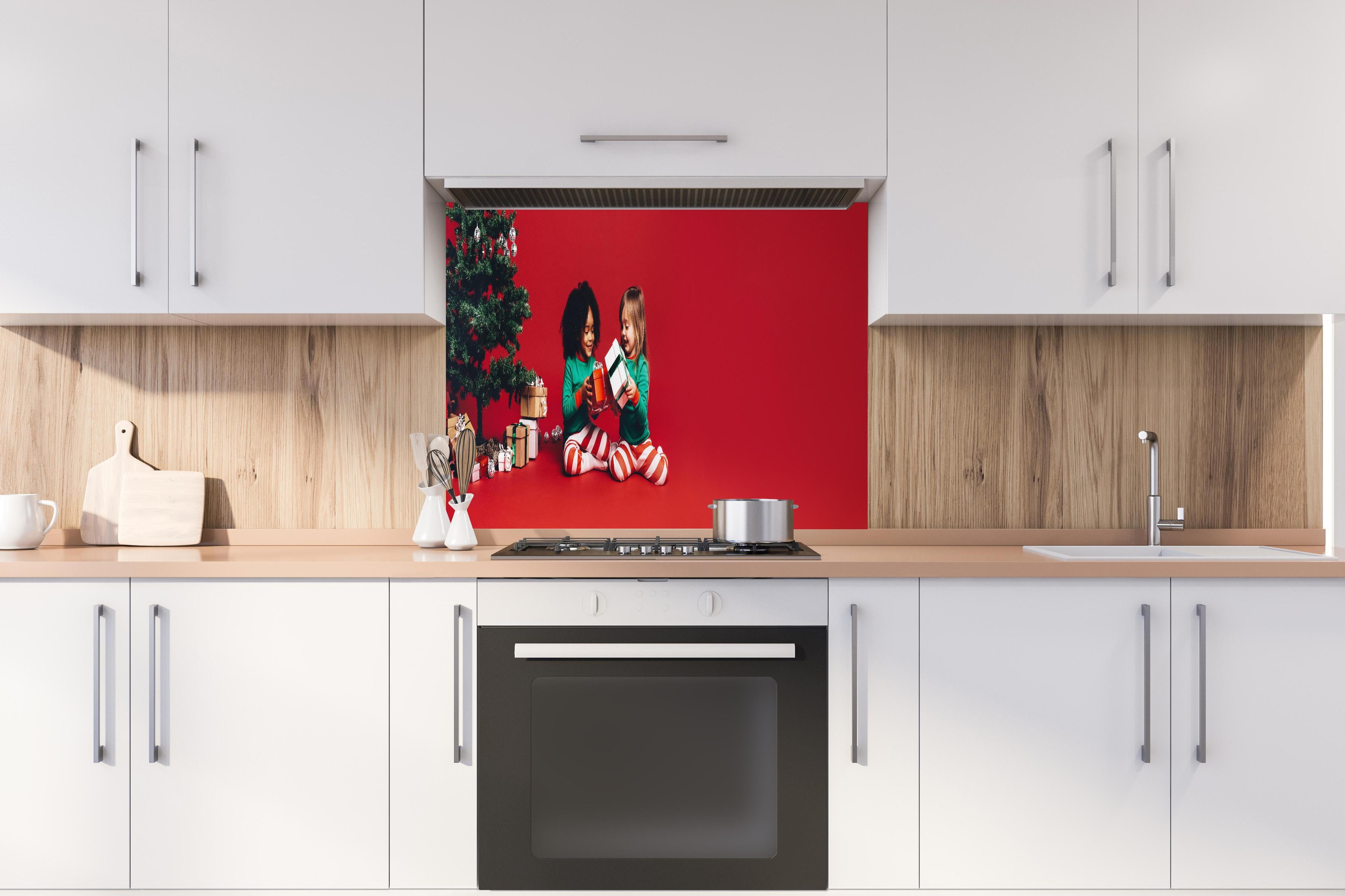 Spritzschutz - Fröhliche Kinder tauschen Weihnachtsgeschenke aus hinter einem Cerankochfeld zwischen Holz-Kochutensilien