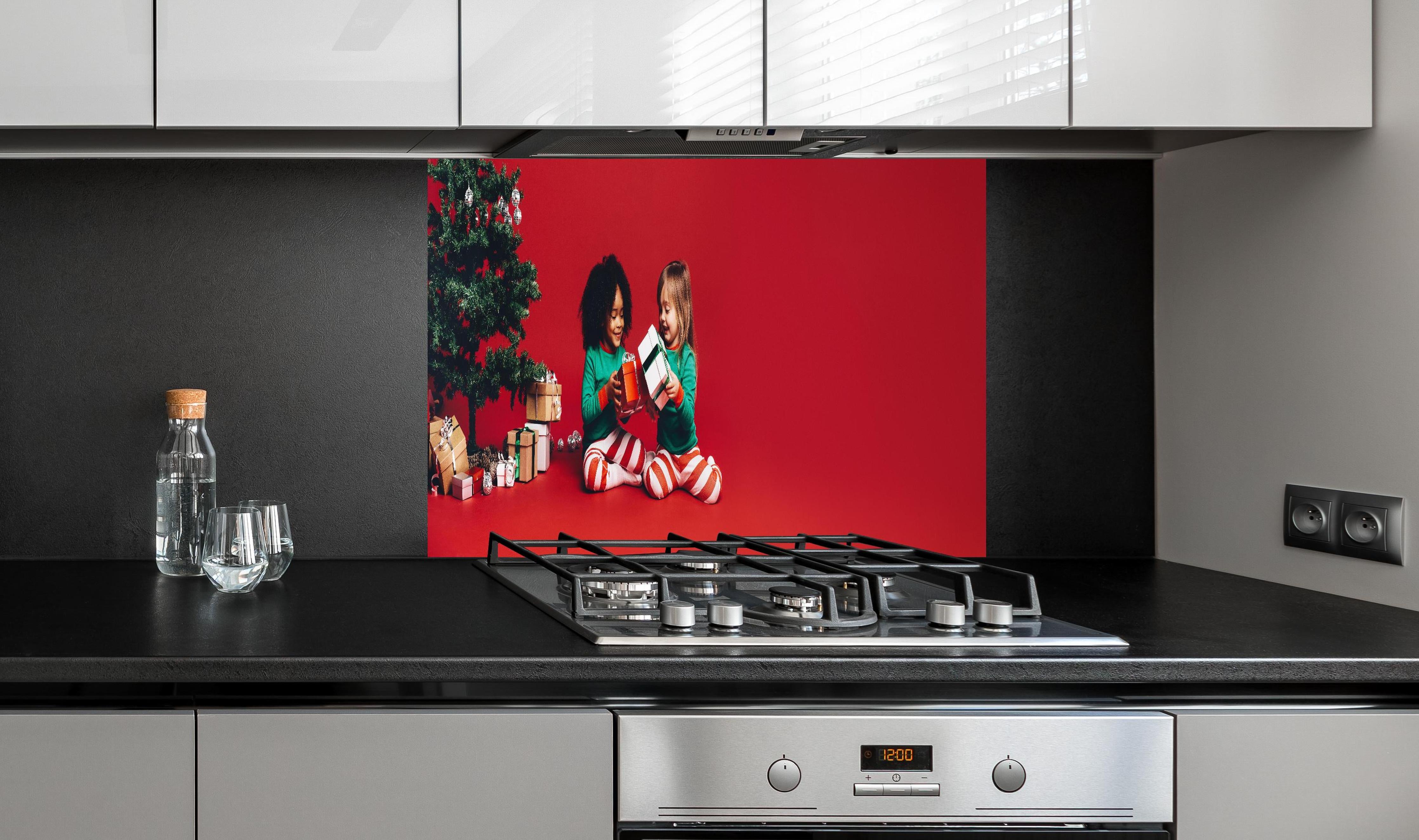 Spritzschutz - Fröhliche Kinder tauschen Weihnachtsgeschenke aus hinter einem Cerankochfeld zwischen Holz-Kochutensilien